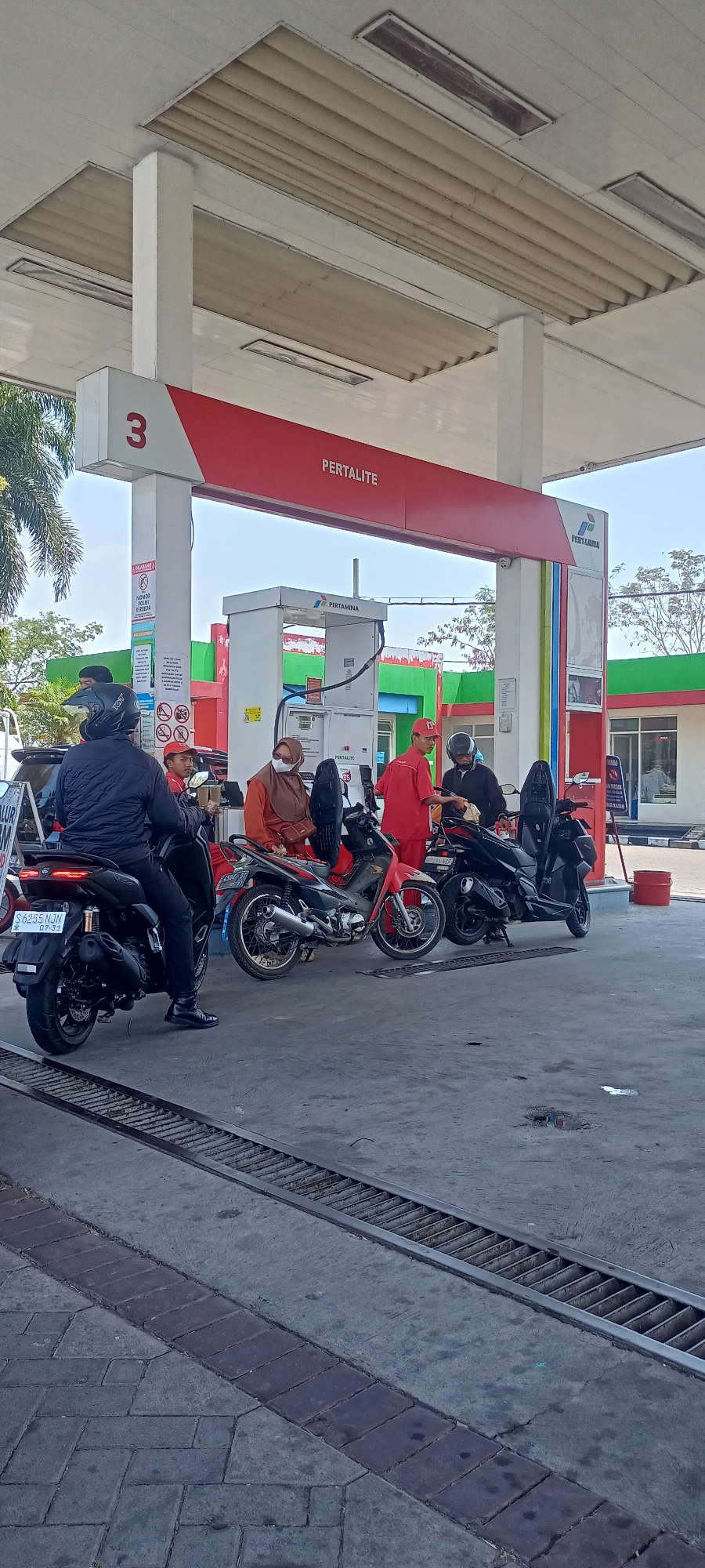 The image depicts a scene at a Pertamina gas station in Sampangagung, Indonesia. Several motorcycles are lined up at a fuel pump. A person wearing a black jacket and helmet is on a black motorcycle in the foreground, facing away from the camera. Next to them is a red motorcycle with a rider. Further back, a person in an orange hijab and a face mask is standing, and a person in a red uniform is interacting with someone on a black motorcycle. Another person in a red uniform is visible on the right. The gas station canopy with the number "3" and "PERTALITE" signage is prominently displayed. The environment appears to be an outdoor urban setting with a paved ground and some vegetation visible in the background.