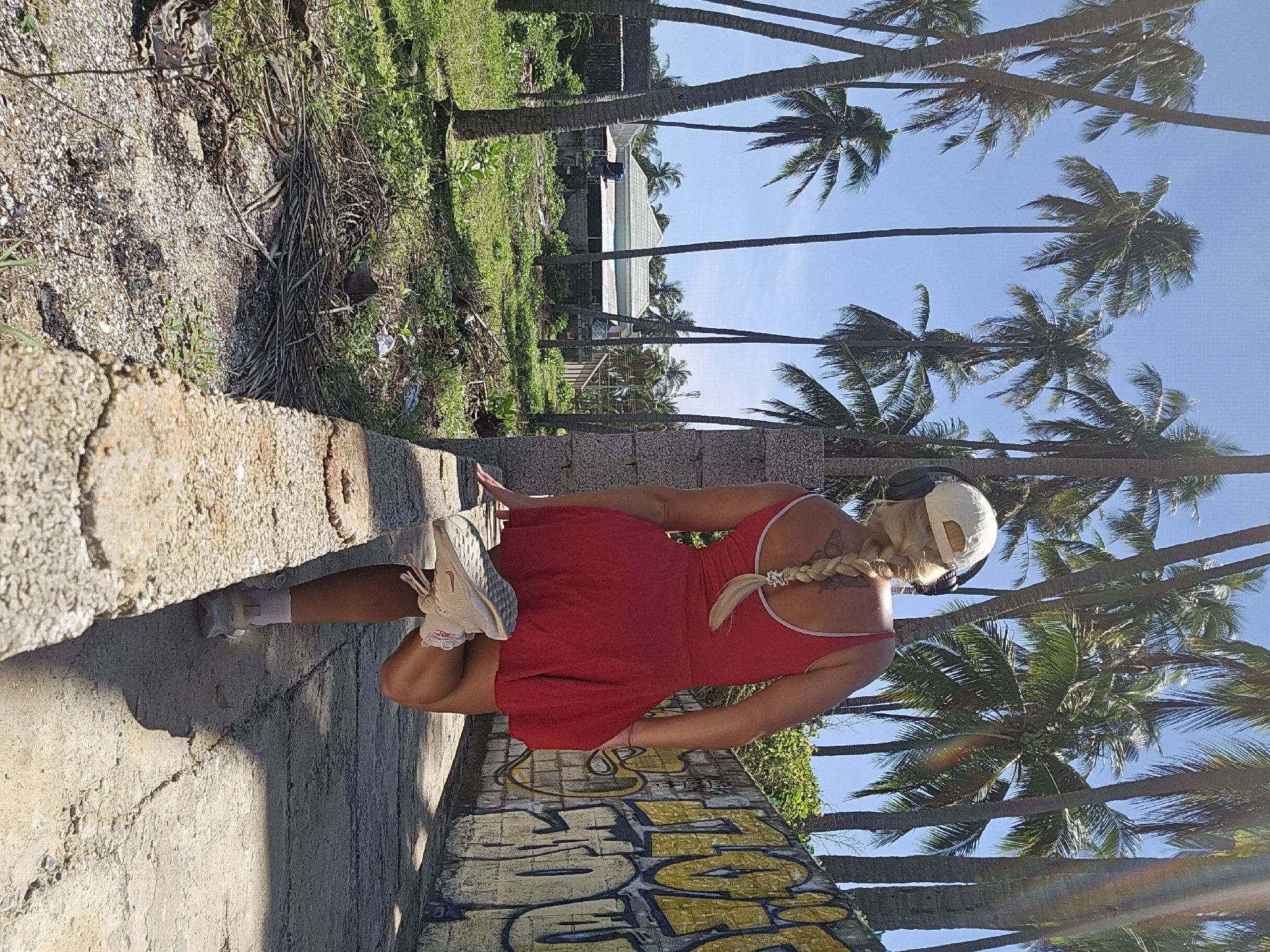 A blonde woman in a red dress and white sneakers is captured in a dynamic pose, facing away from the camera with her right leg extended backwards. She is wearing headphones and a white baseball cap. She is standing on a low concrete wall, with a second concrete wall on her right, adorned with yellow graffiti. The environment is tropical, featuring numerous tall palm trees and lush green vegetation. In the background, a simple house with a tiled roof is visible. The ground is a mix of gravel and dirt, with some debris scattered around. The sky is clear and blue. The lighting suggests it is daytime, with bright sunlight casting shadows. This scene is set in Phu Thuy Ward, Vietnam.