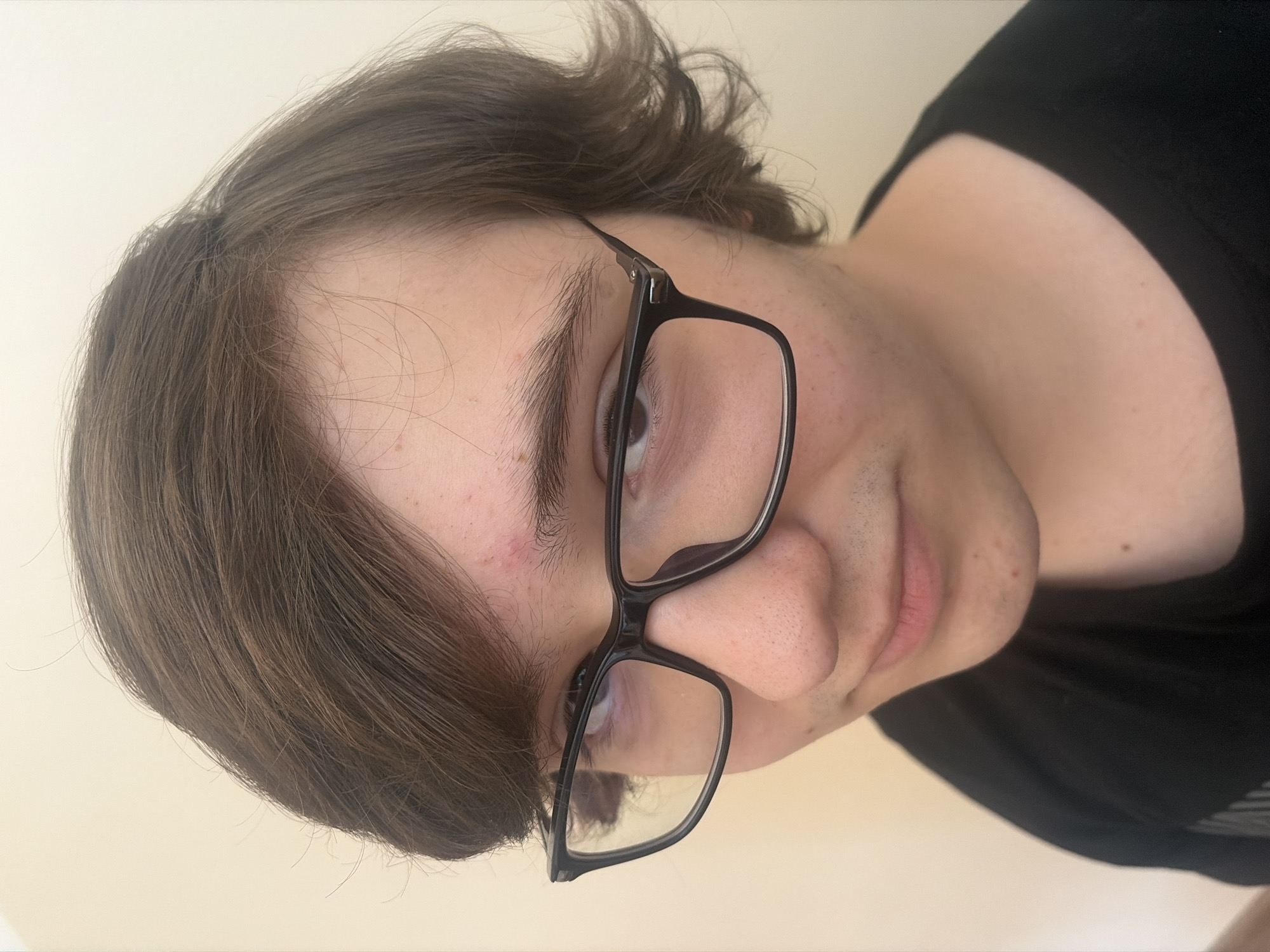 A medium shot shows a young person wearing black-rimmed glasses and a black t-shirt. The person has brown hair that falls across their forehead and partially obscures their right eye. Their left eye is visible, looking towards the viewer. The background is a plain, light-colored wall. No specific location in Sofia, Bulgaria, can be identified from the image.