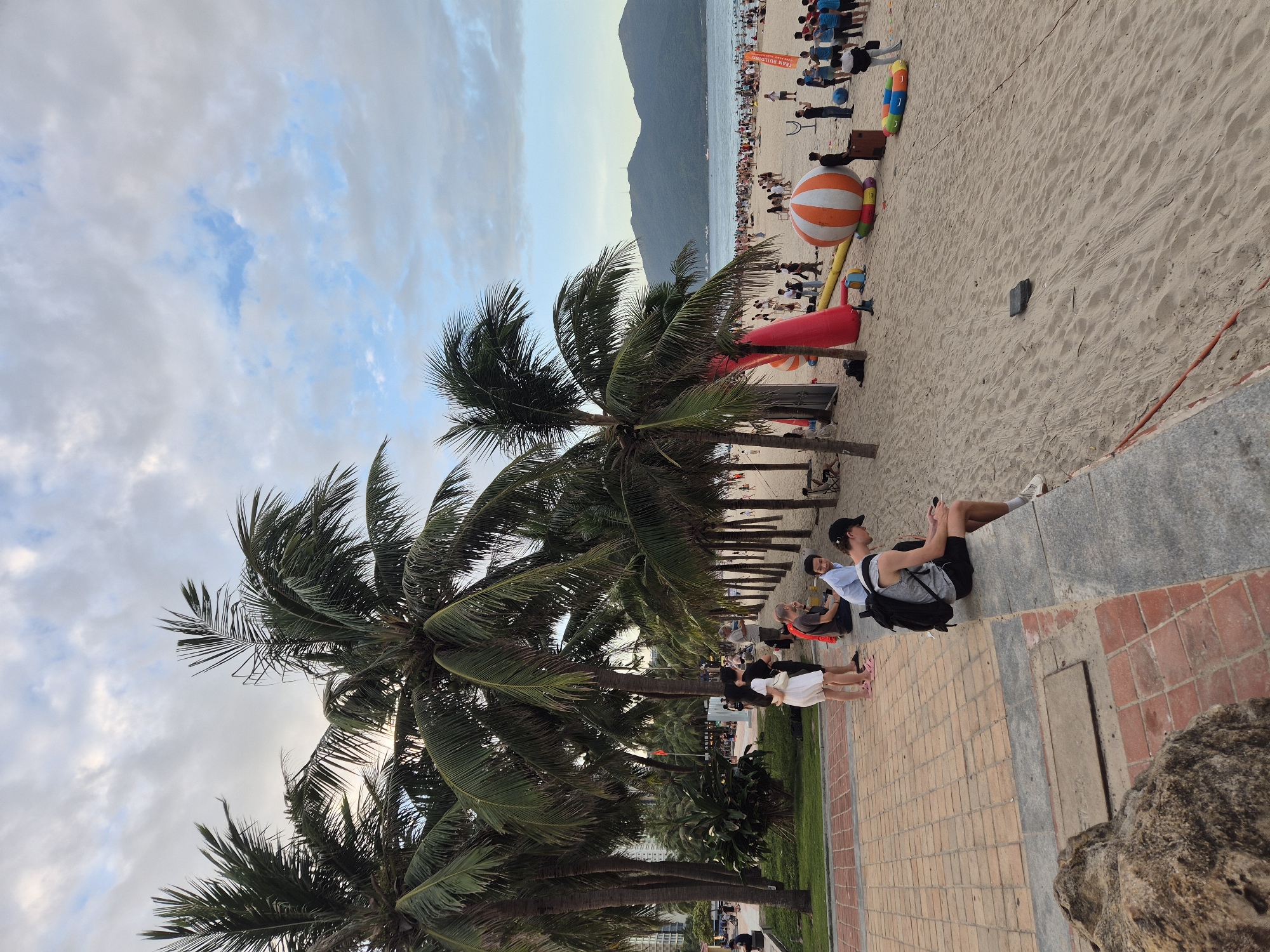The image depicts a bustling beach scene in Da Nang, Vietnam, likely during the late afternoon or early evening. A wide, sandy beach is populated with numerous people, some wading in the water in the distance, while others gather on the shore. Several palm trees line the edge of the beach, providing shade.
In the foreground, a paved walkway runs alongside the beach, bordered by a low wall. Several individuals are seated or standing on this walkway. One young man in a grey tank top and black shorts is sitting on the wall, engrossed in his phone. A couple, a woman in a white dress and a man in a black t-shirt, stand together nearby. Further down the walkway, other people are also seen sitting and conversing.
The beach itself features various activities and objects. Inflatables, including a large orange and white striped ball and a red tube, are present, suggesting recreational events or play areas. A group of people, many wearing blue shirts, are gathered in the mid-ground, possibly participating in an organized activity. In the background, the sea stretches out towards a mountainous coastline under a cloudy sky. The overall impression is one of a vibrant public space where locals and tourists alike are enjoying leisure time.