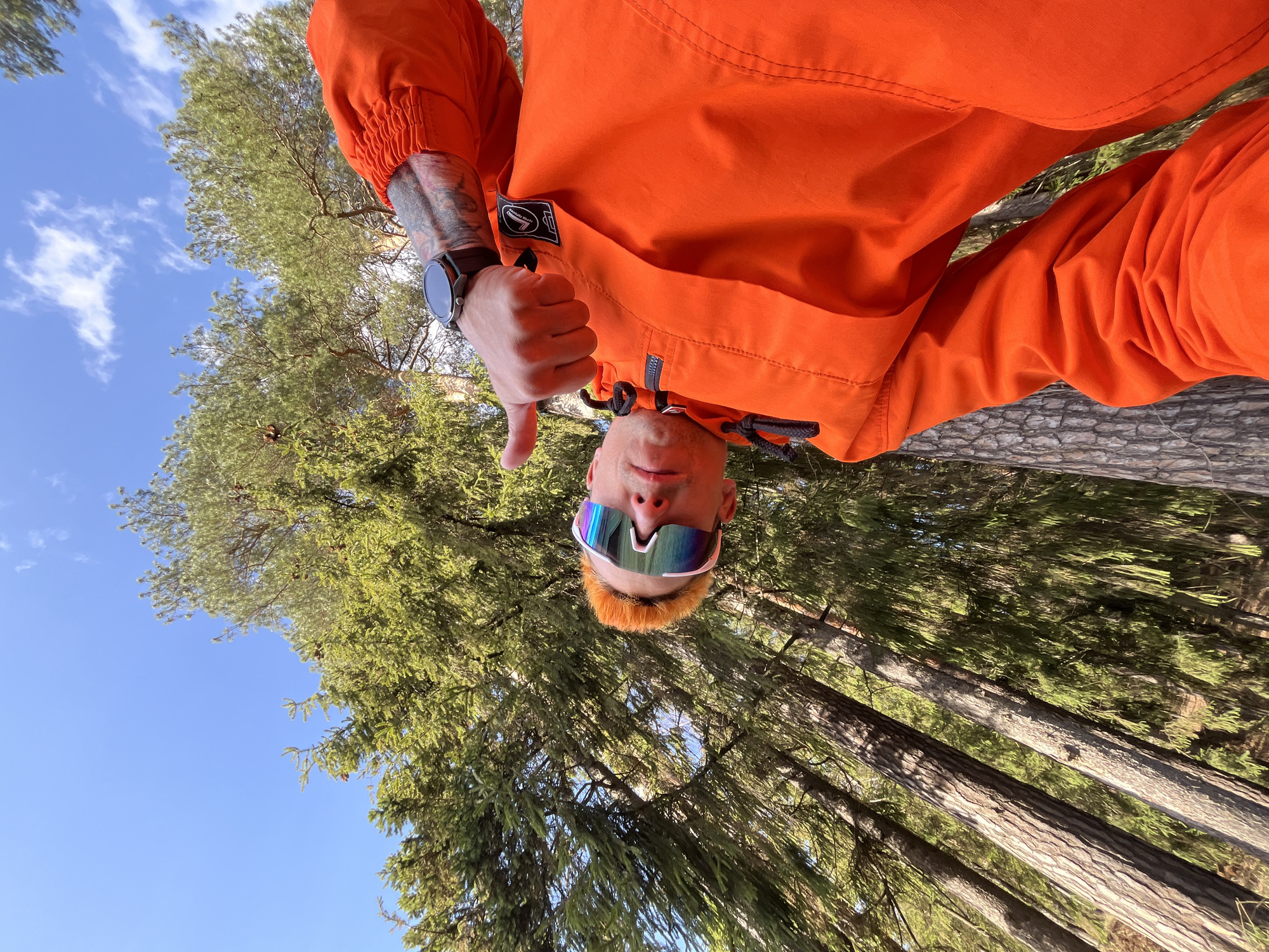 The image, taken from a low angle, shows a man in an orange jacket giving a thumbs down gesture. He is wearing sunglasses and has orange hair. His left wrist has a dark-colored smartwatch. The background consists of tall trees and a blue sky. The setting appears to be a forest, possibly within or near the city of Chaikovsky, Russia. The man seems to be standing or sitting on a tree trunk or log.