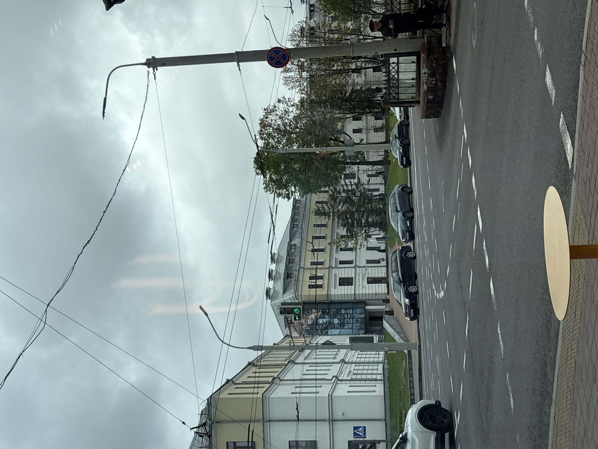 The image depicts a street scene in Minsk, Belarus, taken from a slightly elevated perspective.
The scene showcases a multilane road with parked cars on the right side. A pedestrian is standing near a raised flower bed beside the road. Street lamps and traffic signals are present. A no-stopping sign is visible on one of the poles. Several buildings, varying in architectural style, flank the road. One of the buildings features a modern glass facade. The sky is overcast. A table on the edge of the photo is visible in the foreground.