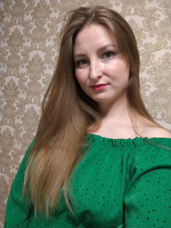 A medium close-up, indoor portrait captures a young woman looking directly at the camera. She has long, straight to slightly wavy, light brown or caramel-colored hair that is parted slightly off-center to her left and flows over her shoulders and chest. Her face is oval-shaped with fair skin and appears to have a subtle, natural makeup application. Her eyes are light-colored, possibly green or hazel, and she has visible eyelashes. Her lips are full and adorned with a pink or fuchsia lipstick. She has a neutral to slightly pleasant expression, with a hint of a gentle smile.
She is wearing an off-the-shoulder top in a vibrant emerald green color. The top features small, irregular black polka dots or speckles scattered across the fabric. The neckline is elasticized and ruffled, exposing her right shoulder and the upper part of her chest. The top also has smocked detailing visible at the bottom of the frame, around what appears to be the waist or hemline.
The background consists of a wall covered in ornate wallpaper. The wallpaper features a classic damask-like pattern with intricate floral and foliate motifs in muted gold, beige, and light brown tones, creating a textured and elegant backdrop. The lighting in the image suggests an indoor setting, likely natural or soft artificial light, illuminating her face evenly. No text is visible in the image.