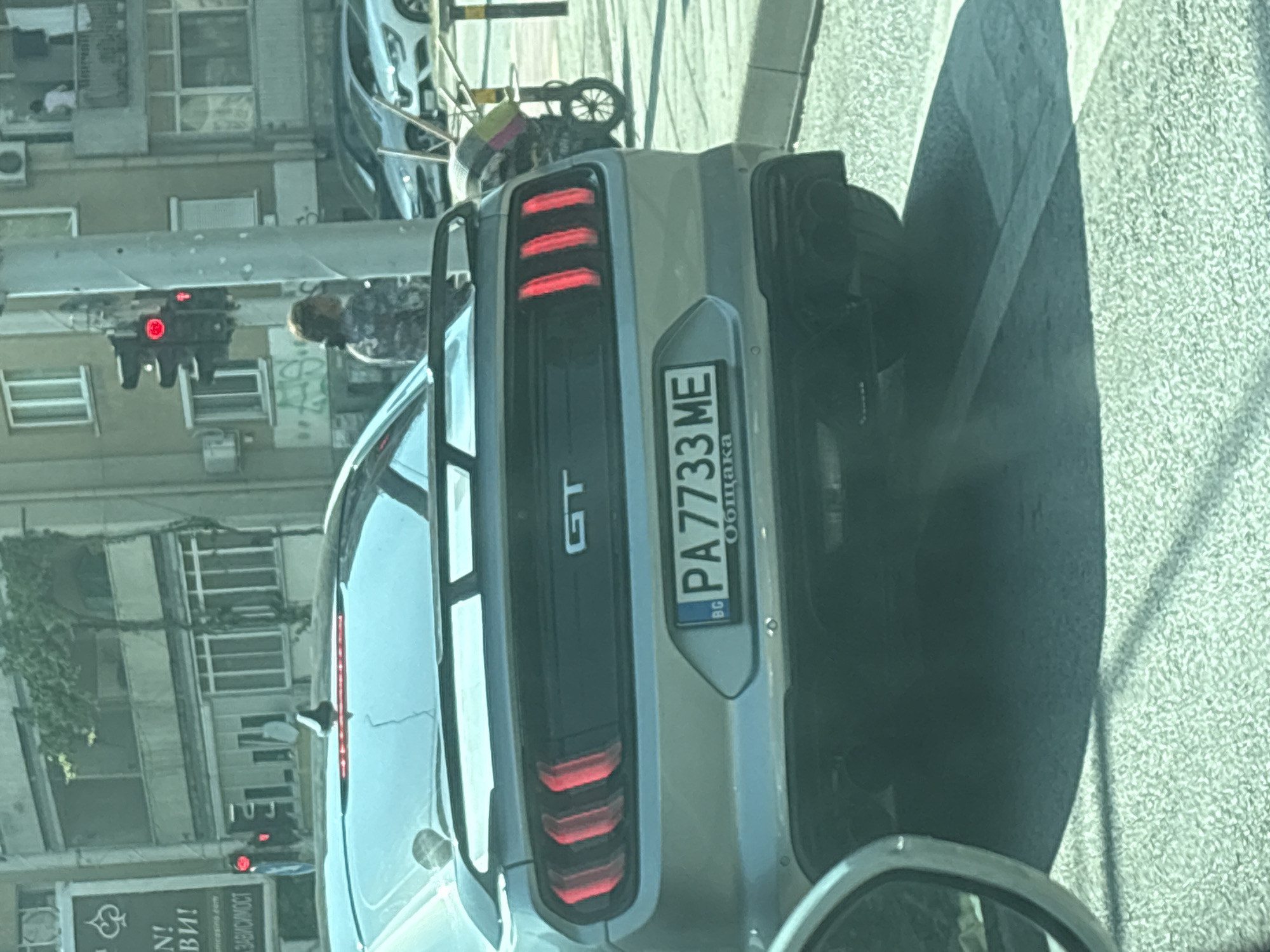 The image captures the rear view of a silver-grey Ford Mustang GT coupe stopped at a red traffic light on a city street in Plovdiv, Bulgaria. The car's license plate, prominently displayed, reads "PA 7733 ME" with "BG" indicating Bulgaria and the word "Община" (Municipality) below the numbers. The Mustang features a black rear spoiler and distinctive vertical triple LED taillights illuminated red. Dual exhaust pipes are visible on the lower right of the rear bumper.
To the left, a partially visible white sign with Cyrillic text shows "N!", "ВИ!", and "ЗАВИСИМОСТ". In the middle ground, a woman with dark, pulled-back hair, wearing a patterned top, stands near a pole with the red traffic light. The traffic light itself is a standard three-section vertical signal, with the top red light illuminated.
The background consists of multi-story residential buildings with balconies and windows. One building has green vines growing on its facade, and another shows a person standing on a balcony. Air conditioning units are mounted on the exterior walls of some buildings. Several other cars are parked or stopped further down the street. On the right side of the street, a small, wheeled cart or stroller with a multi-colored canopy is positioned near the curb. The ground is paved with asphalt, showing light and shadow patterns, suggesting a sunny day. The perspective is from inside another vehicle, with part of its side mirror visible in the lower left foreground.