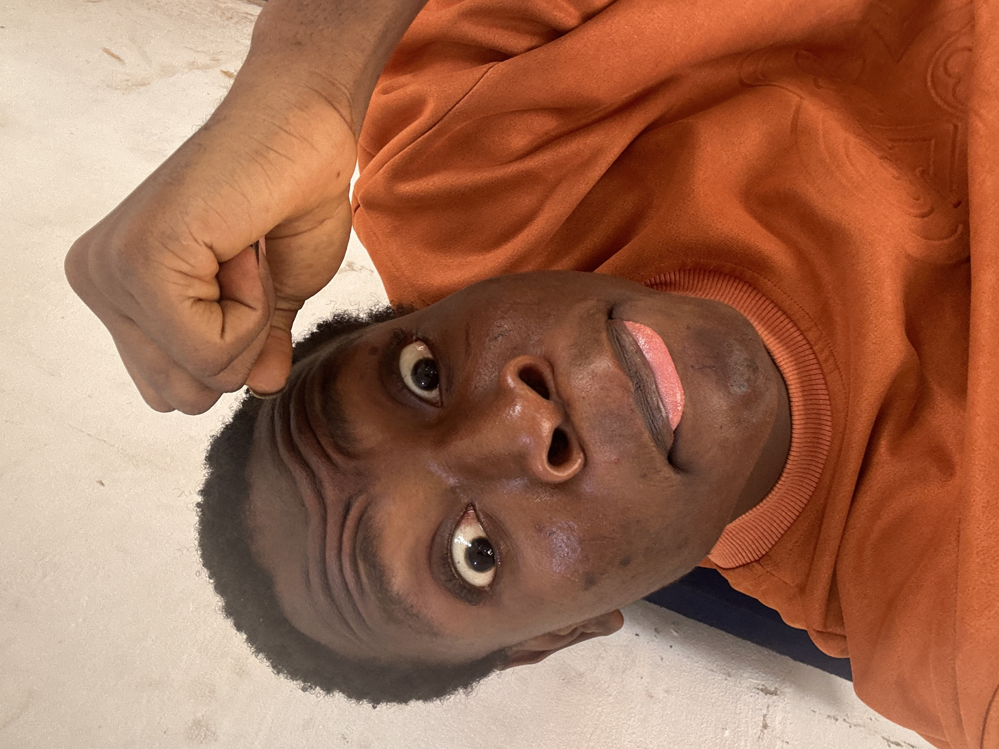 A close-up shot, oriented upside down, depicts a person with dark skin wearing an orange sweater. The person's face is prominent, with visible wrinkles on the forehead. One hand, with a clenched fist, is positioned near the head. The background appears to be a light-colored surface. The city and specific location are unknown.