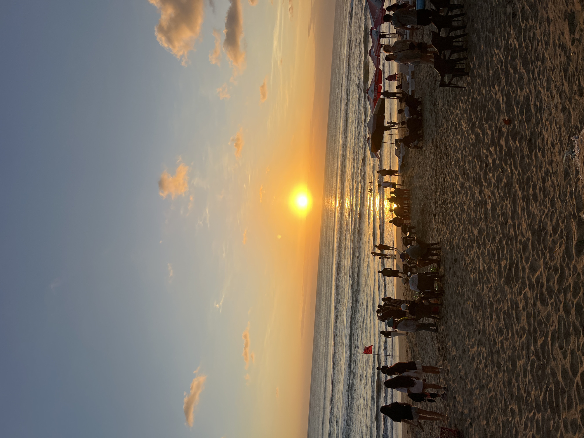 The image depicts a sunset scene at Kuta Beach, Bali, Indonesia. The sun is setting over the ocean, casting a warm golden glow across the sky and water. Waves are gently rolling onto the shore. The beach is populated with people, some standing and others sitting on chairs or lounging under umbrellas. A red flag is visible on the beach. The sand is textured with footprints and ripples. The sky is a gradient of blue and orange, with a few scattered clouds.