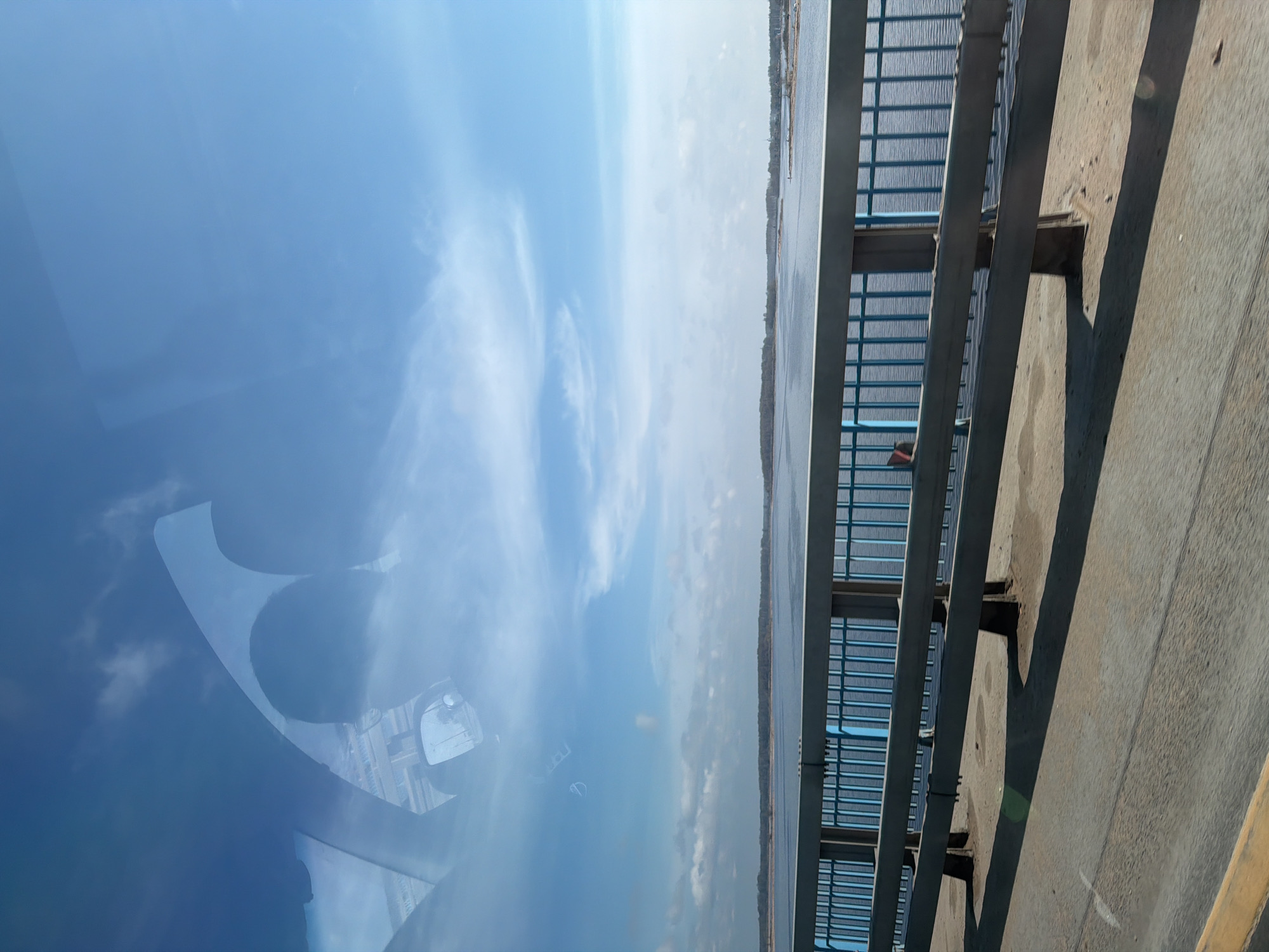 The image appears to be taken from inside a vehicle driving across a bridge. The sky and clouds are reflected in the windshield. A body of water is visible next to the bridge, with trees lining the shore. The bridge has a metal railing. The location is Bezborodovo, Russia.