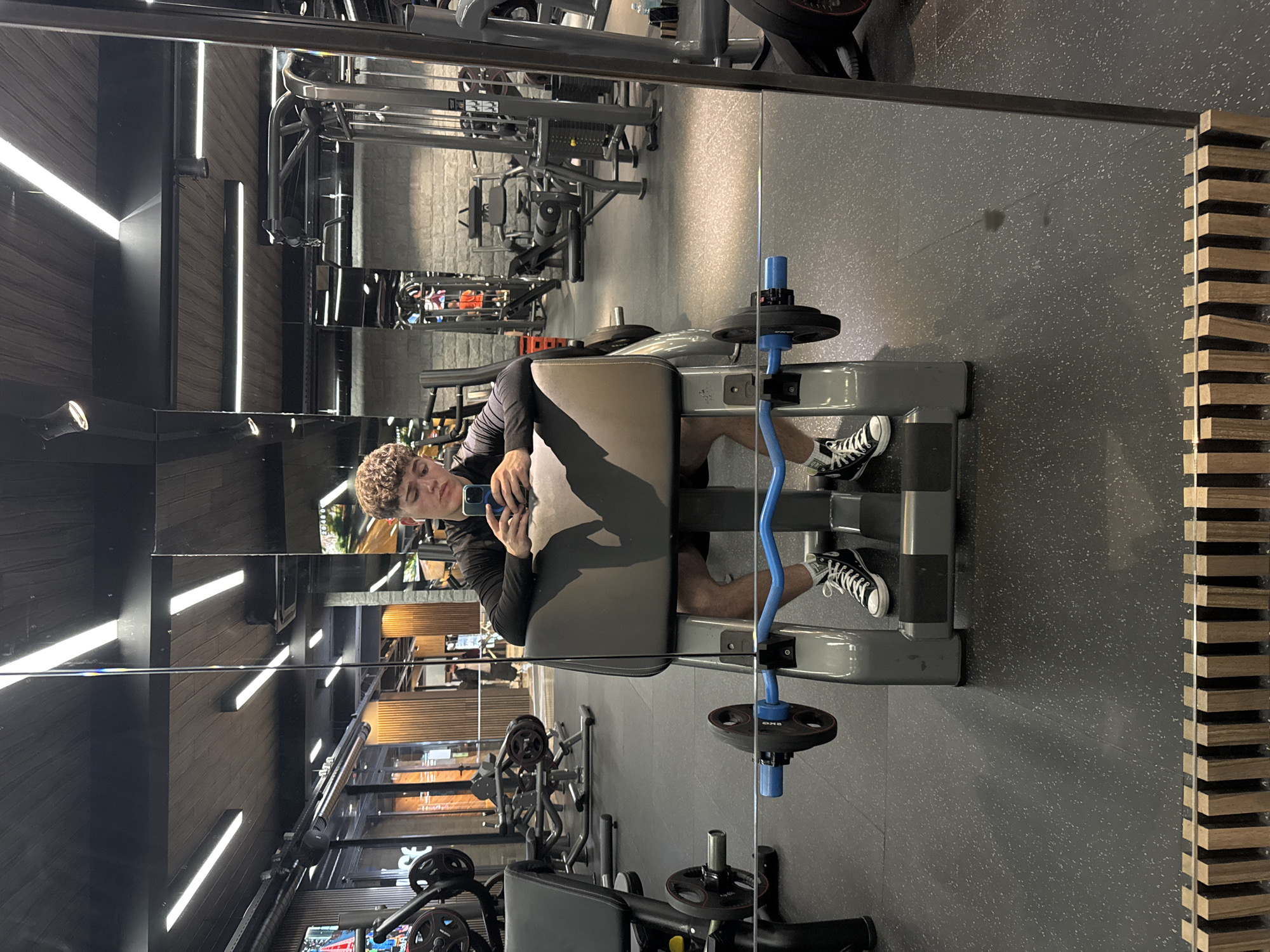 This image is a mirror selfie taken by a young man inside a modern gym. The man, with light brown curly hair, is wearing a dark long-sleeved shirt and black and white sneakers, which appear to be Converse Chuck Taylors. He is seated on a grey gym machine, possibly for leg exercises, and is holding a blue smartphone in his hands, looking towards the camera (his phone).
The large mirror reflects the gym's interior, showing a well-lit space equipped with various fitness machines and weights. A blue barbell with black weight plates is visible near the man's feet on the machine. The gym floor is dark grey with white speckles, typical of rubber gym flooring. The walls feature dark wood paneling, and the ceiling and walls are illuminated by bright, linear LED lights, creating a contemporary atmosphere. A decorative wooden slatted structure is visible on the right side of the reflected background.
The activity depicted is the young man taking a mirror selfie, likely during a break or before commencing his workout. His expression is neutral as he focuses on his phone. No specific time of day can be determined due to the indoor setting and artificial lighting. No legible text is visible on any objects or clothing within the frame. The location context provided is Sofia, Bulgaria.