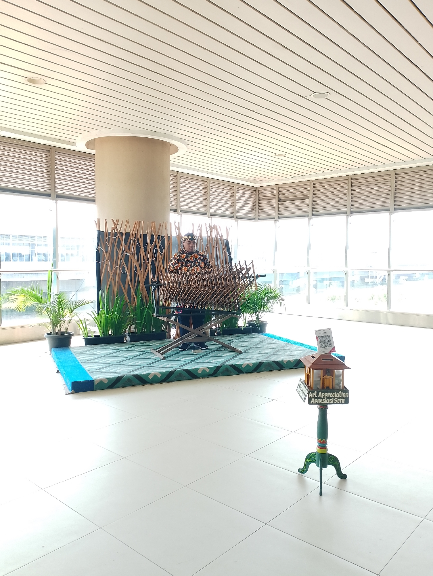 The image depicts a brightly lit, modern interior space with large windows. In the foreground, a decorative stand with a miniature house structure is placed on the tiled floor. The stand is green with colorful accents and has a sign that reads "Art Appreciation Apresiasi Seni". To the left, a green and blue patterned mat is laid out, with a person standing behind a musical instrument that resembles a gamelan. The person is wearing a traditional outfit and a head covering. Several potted plants are arranged around the instrument. The background features a large cylindrical pillar and a wall with horizontal louvers. The overall scene suggests an indoor cultural exhibition or performance space.