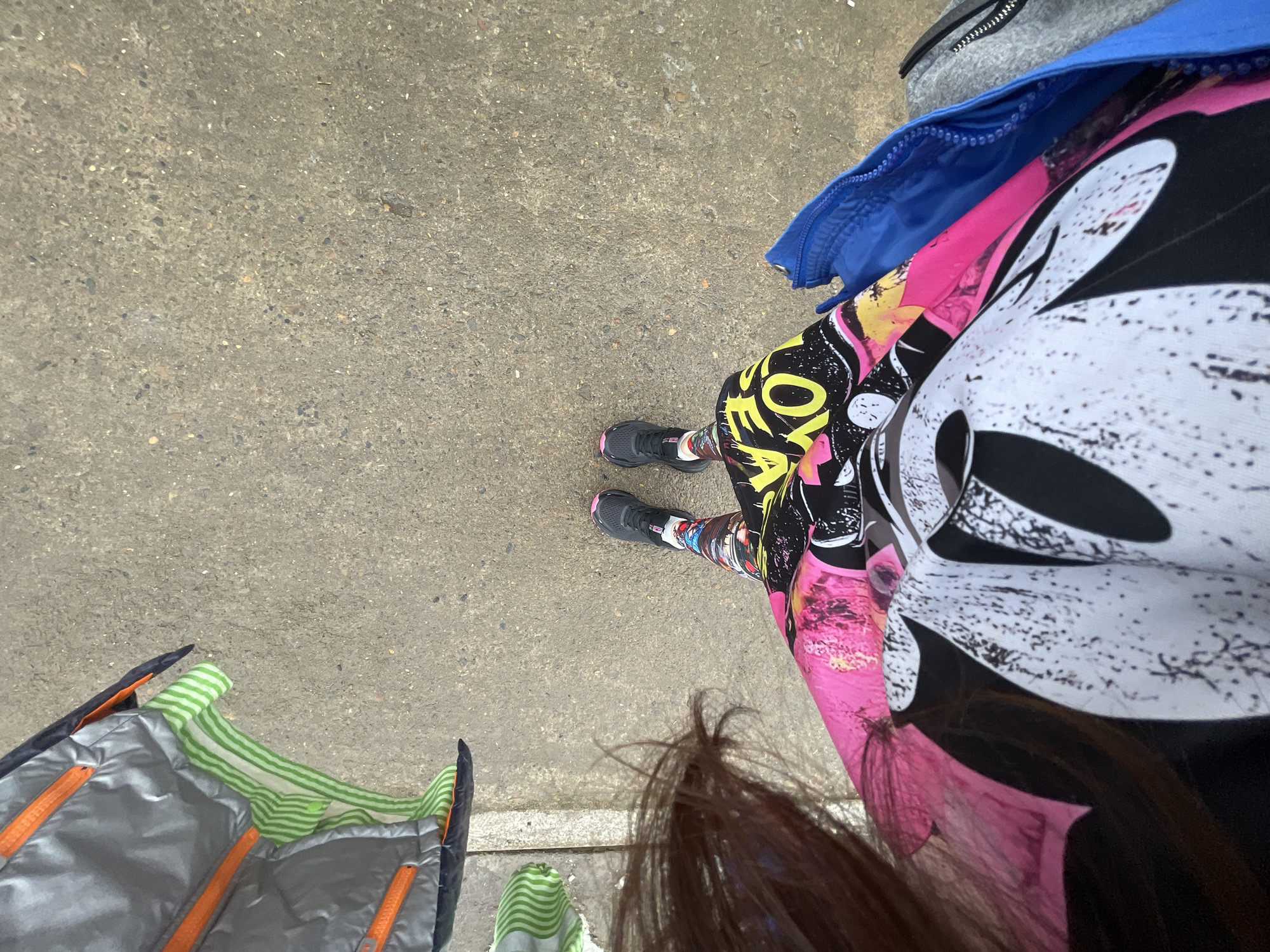 An aerial perspective captures a section of a concrete or asphalt surface in Chaikovsky, Russia, featuring a standing individual and several objects. The individual, observed from the mid-torso downwards, wears a vibrant pink garment layered over a black top with a prominent white, distressed graphic resembling Mickey Mouse. Below the waist, multi-colored leggings with abstract and textual patterns, including partial yellow text reading "OY," are visible. The individual's footwear consists of black athletic shoes accented with pink on the soles. To the right of the individual, a blue jacket with an exposed zipper is present, partially obscuring a grey garment. To the left, a grey, possibly metallic or padded, rectangular object with prominent orange zippers rests on the ground. Adjacent to this object, two green and white striped socks are visible, one partially unfolded, the other folded. A white painted line runs horizontally across the concrete surface beneath the grey object and socks.