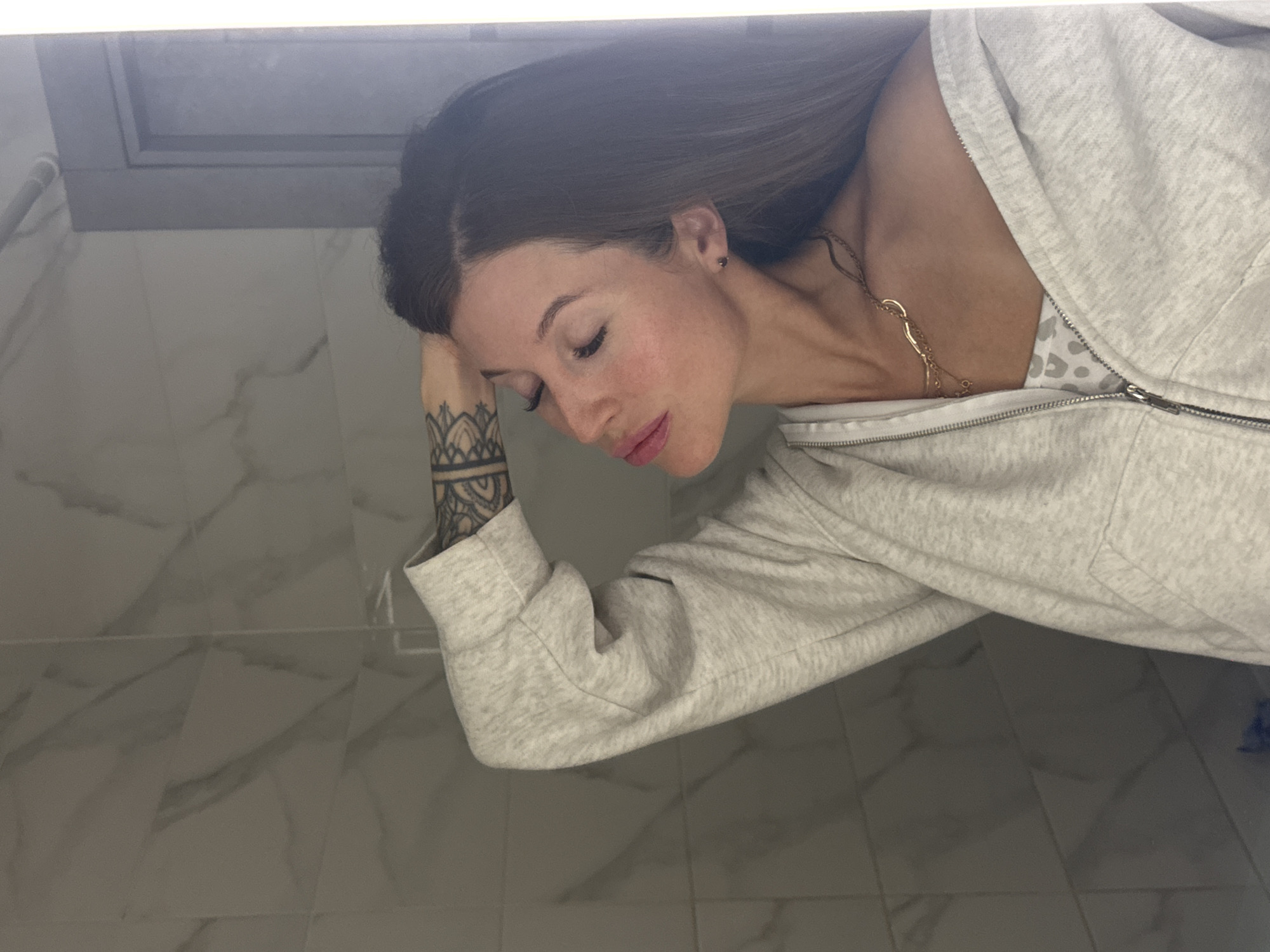 A fair-skinned woman with long brown hair rests her head on her left arm, which is tattooed. She wears a light grey zip-up hoodie over a spotted garment and a gold necklace. The background is a marble-tiled wall and potentially a vanity area. There is no indication of Chaikovsky, Russia, or any other specific location.