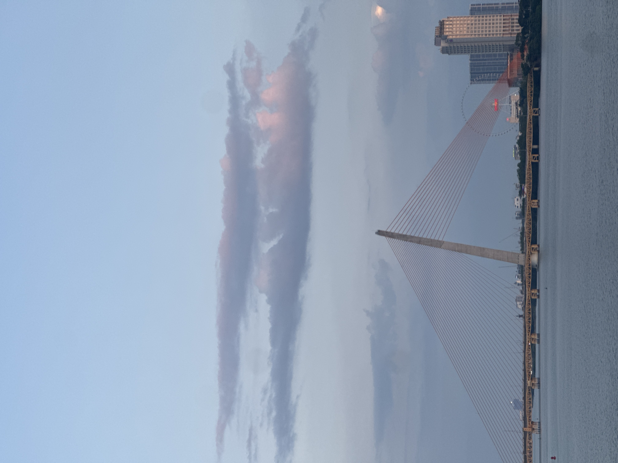 The image depicts the Trần Thị Lý Bridge in Đà Nẵng, Vietnam, at dusk. The bridge is a cable-stayed bridge over the Hàn River. Its tall concrete pylon rises skyward, with red cables fanning out to support the roadway. A smaller, traditional girder bridge is seen adjacent to the cable-stayed structure. In the background, the illuminated Sun Wheel and city buildings can be seen on the riverbank. The sky is filled with blue and grey clouds, tinted pink by the setting sun. The river water is calm and reflects the sky.