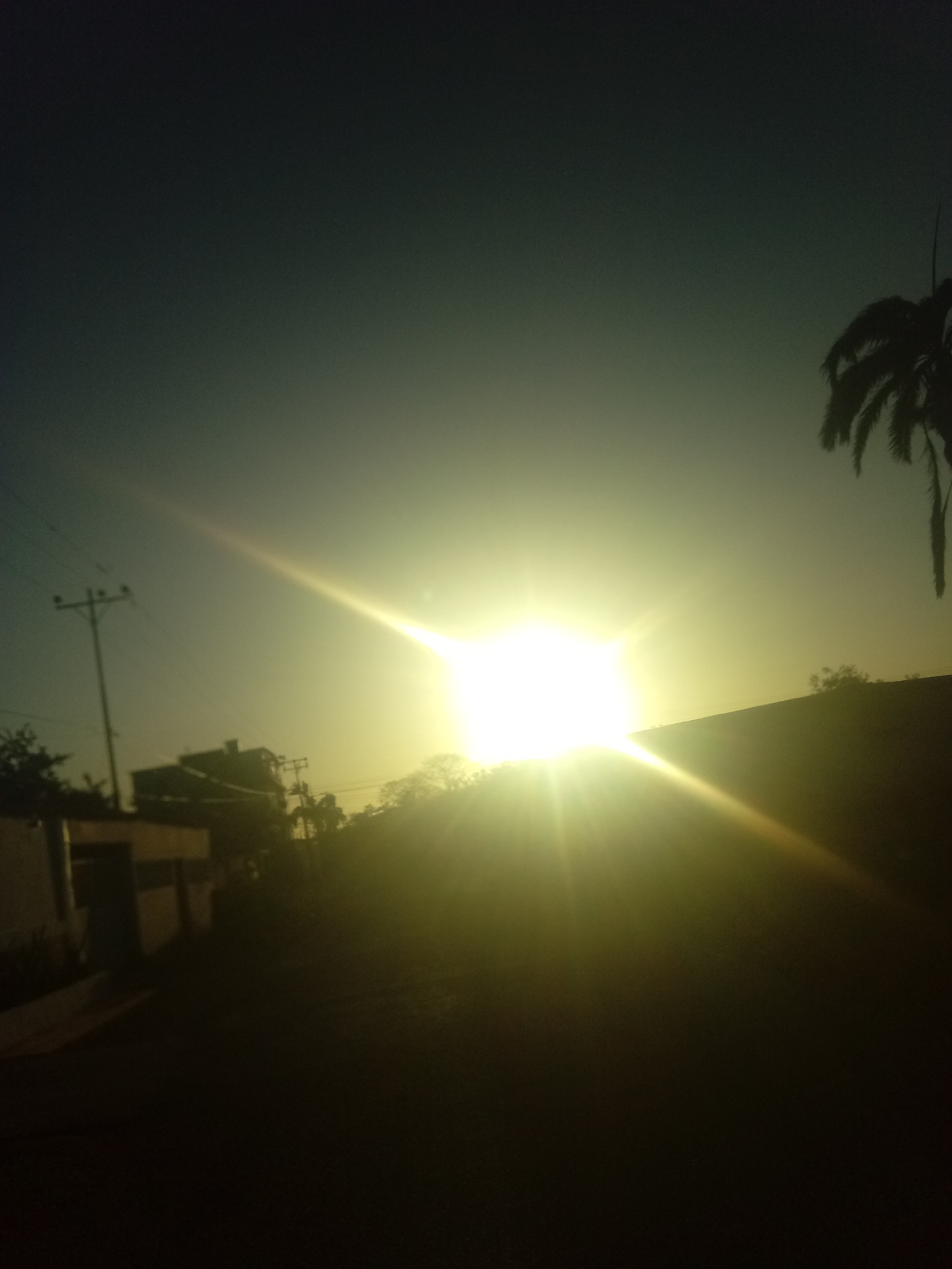 The image shows a bright sunrise or sunset in Juan Griego, Venezuela. The sun is the dominant feature, positioned in the center of the frame, emitting strong rays of light. The foreground is dark, suggesting it's a road or pathway. In the midground, there's a silhouette of a building or houses with utility poles and power lines nearby. A palm tree can be seen on the right side of the frame. The sky transitions from a darker hue at the top to a lighter, yellowish tone near the horizon where the sun is positioned.