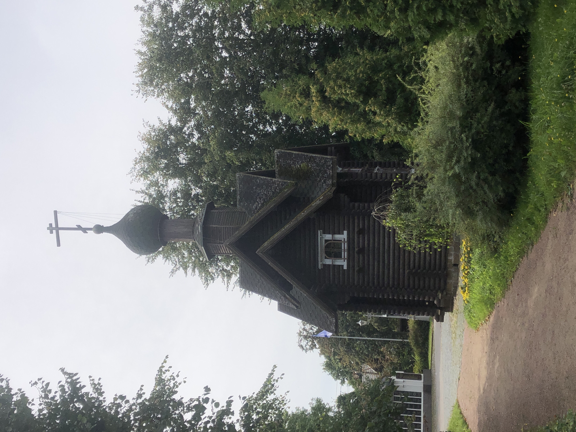The image depicts a dark wooden chapel with a green, onion-shaped dome topped with a cross, situated amidst lush greenery. A dirt path leads towards the chapel, which has a small, square window with a decorative white frame. Behind the chapel, a flagpole with a blue and white flag is visible. The overall scene suggests an outdoor setting, possibly a park or a historical site, under an overcast sky. The architectural style of the chapel, characterized by its log construction and domed roof, is consistent with traditional Russian Orthodox churches. The presence of mature trees and dense foliage indicates a temperate climate. The chapel appears to be a significant landmark within its surroundings.