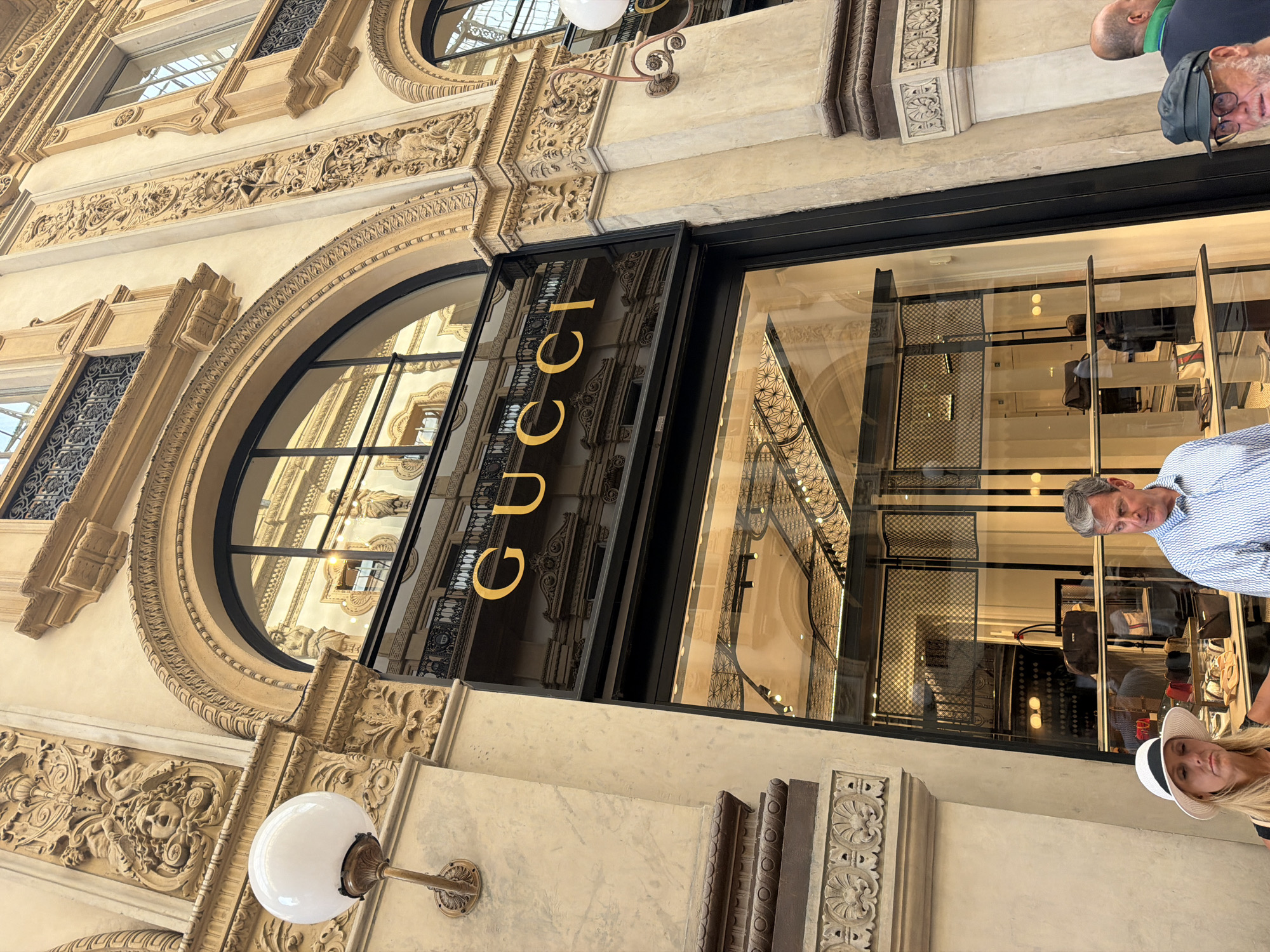 The image captures the exterior of a Gucci store situated in Milan, Italy. The architectural design of the building exhibits ornate, classical features. The Gucci logo is prominently displayed above the entrance. Reflections are visible in the store's glass windows, hinting at an elaborate interior design and merchandise. The foreground shows a person wearing a hat, while another person is partially visible, suggesting the scene is a public space with people passing by or observing the store.