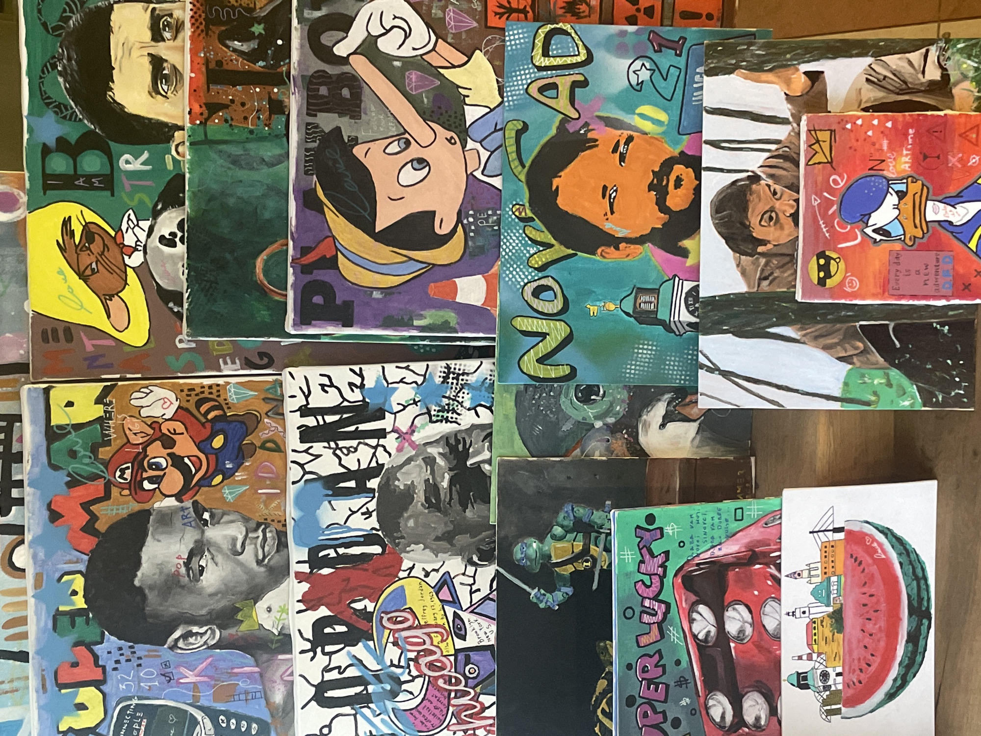 The image presents a collection of small, colorful paintings laid out on a wooden surface. The paintings depict various subjects, including portraits of people like Bob Marley, cartoon characters such as Mickey Mouse, Pinocchio, Donald Duck, Mario, and Leonardo from Teenage Mutant Ninja Turtles. A painting shows Novi Sad, Serbia through a watermelon in half. There are also paintings with text and graffiti-like designs. The style of the paintings appears to be pop art or street art. The environment is a close-up view, focusing entirely on the display of the artwork.