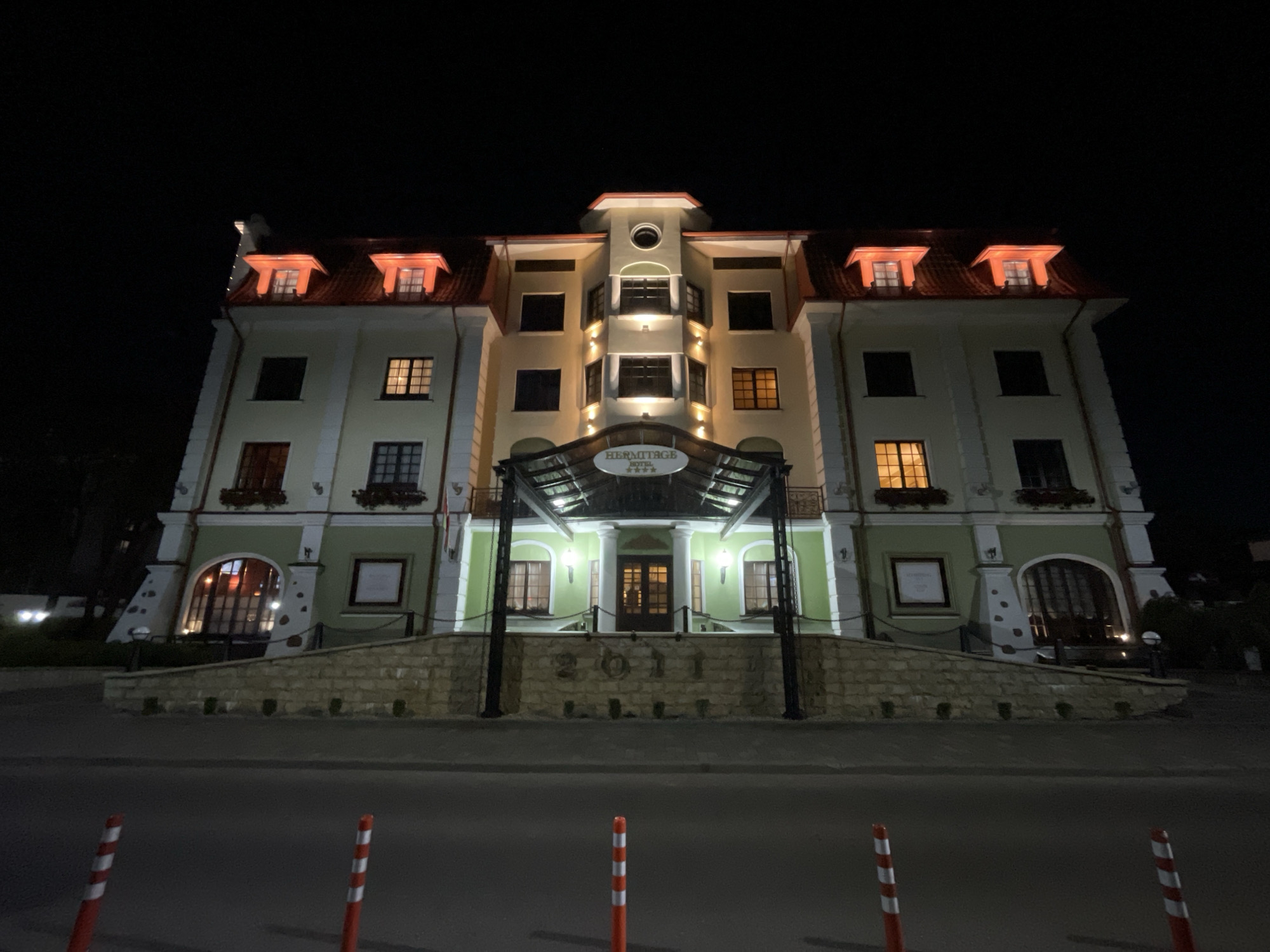 This is a night-time, low-angle, wide shot of the Hermitage Hotel in Brest, Belarus. The hotel is a light yellow and green building with a red tiled roof. It has several dormer windows lit from within, casting an orange glow. The entrance is under a covered walkway supported by dark pillars. Above the entrance, a sign reads "HERMITAGE HOTEL ****". Below the entrance, a stone wall with the number "2011" carved into it is visible. The area in front of the hotel appears to be a road, with red and white striped bollards in the foreground. The sky is completely black, indicating the photo was taken after dark. There are no people visible in the image.