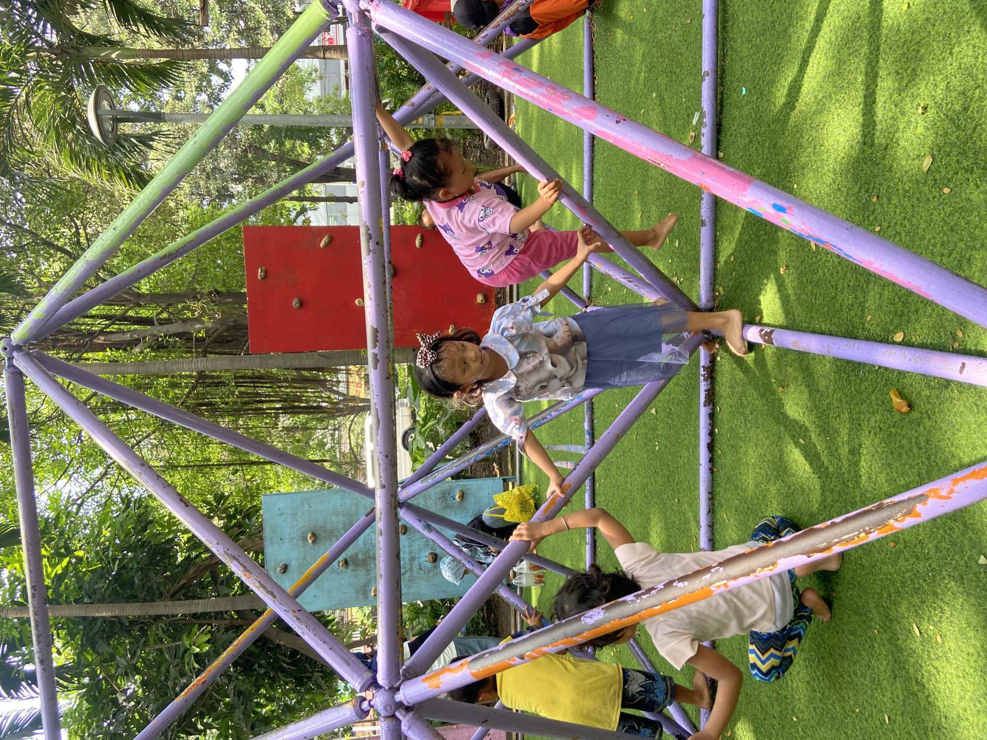 This image captures a vibrant scene of several young children playing on a geometric climbing structure at an outdoor playground in Surabaya, Indonesia. The structure consists of numerous purple-painted metal bars, some with patches of green, pink, and orange paint, forming a large dome or pyramid shape. The ground is covered with bright green artificial turf.
In the upper section of the structure, a young girl in a pink patterned t-shirt and pink pants hangs barefoot from a purple bar, actively climbing. Below her, another girl in a light-colored patterned top and a blue skirt stands within the structure, holding onto a bar and looking towards the camera. Other children are partially visible around the base and inside the structure, interacting with the bars. One child in a yellow t-shirt and patterned shorts is seen near the bottom, while another in a white t-shirt and blue/green patterned shorts is on the turf near a bar.
In the background, two climbing walls are visible: a red one with three visible holds and a blue/turquoise one with multiple holds. Lush green foliage and trees indicate a sunny, daytime setting. A yellow bag with the word "TRUE" is faintly visible near the blue climbing wall. The children appear engaged and joyful in their play.