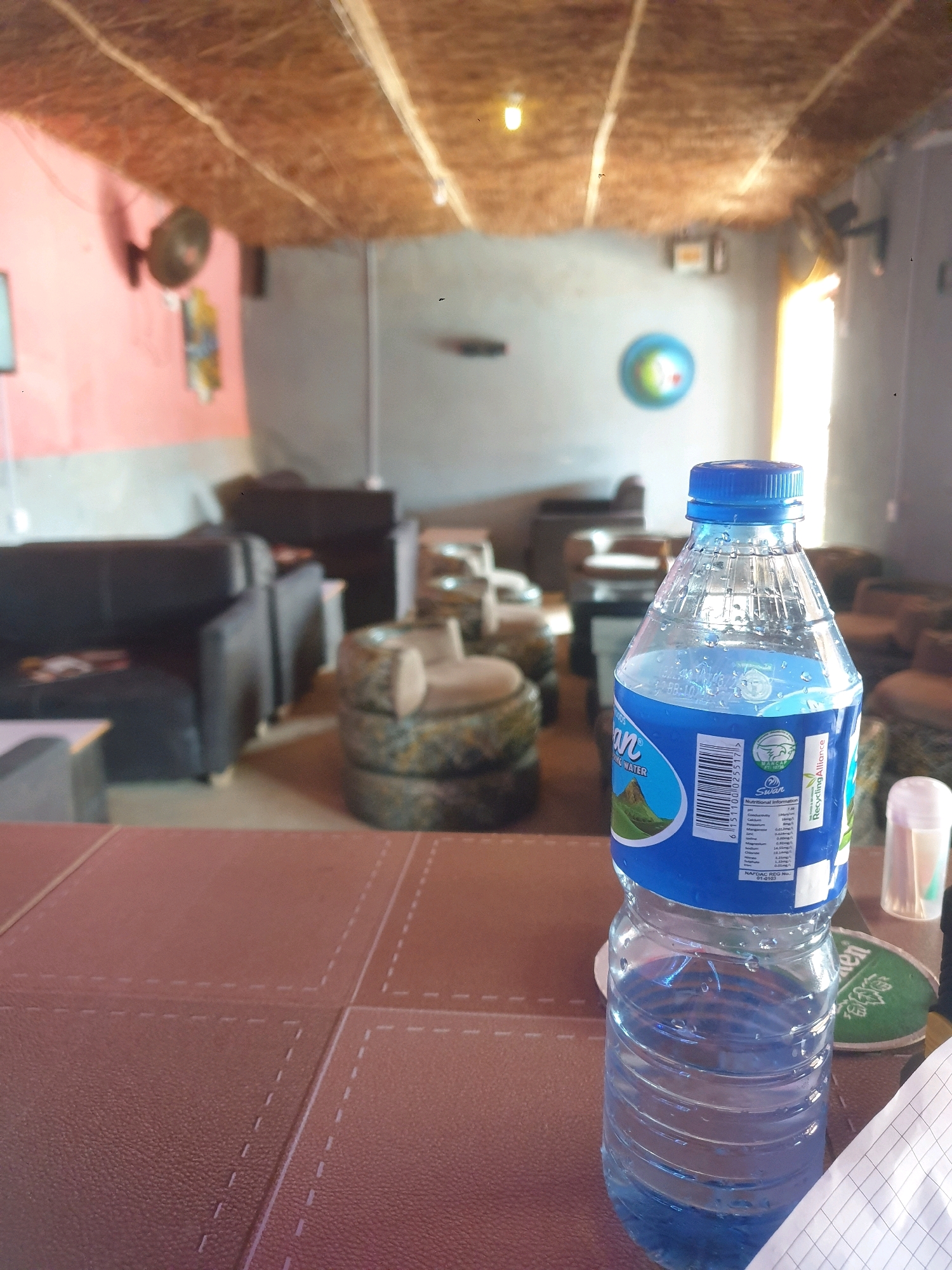 This image depicts a close-up of a plastic bottle of Swan drinking water, filled with blue-tinted water, placed on a brown textured surface. The bottle has a blue cap and a label with text and images, including nutritional information. In the background, out of focus, a room is visible with various seating arrangements, including dark sofas and circular chairs made of stacked tires. The ceiling appears to be made of thatched material. A fan is mounted on a pink wall, and a blue and green circular object is on a gray wall. The lighting suggests an indoor setting, possibly a cafe or lounge. The image is likely taken in Mangu Town, Nigeria.