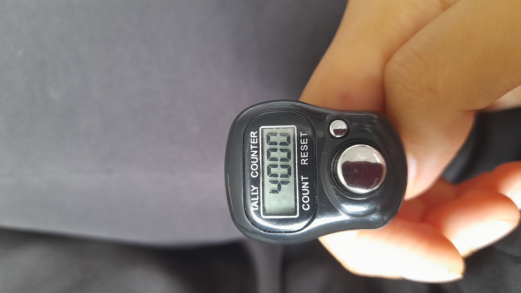 A close-up shot shows a black tally counter being held in a person's hand. The tally counter has a digital display showing the number "4000". Above the display, text reads "TALLY COUNTER", and below the display, text reads "COUNT RESET". A large silver button is visible below the display, and a smaller silver button is to its right. The background is out of focus and appears to be dark gray fabric. The person's fingers are visible in the foreground, holding the tally counter.