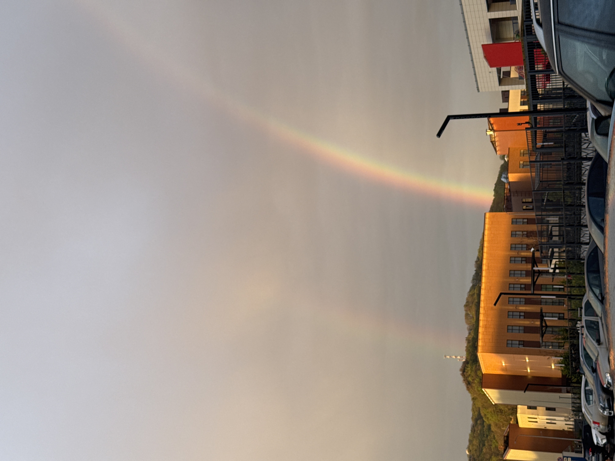 Here is a detailed description of the image:
**Scene:** A landscape-oriented shot captures a rainbow stretching across a cloudy sky. In the lower right portion of the image, there are cars in a parking lot beside a building complex.
**Objects:** A rainbow is visible in the sky. Cars are parked to the right side. Buildings rise in the background, including what appears to be a block of apartments with red highlights and windows. A small antenna is seen against a leafy hill. A lamppost can be seen near the building.
**Environment:** The sky is overcast, giving the scene a somewhat muted color palette. The parking lot and buildings are situated adjacent to some green hills or trees.
**City:** The exact location in Sochi, Russia is unknown.