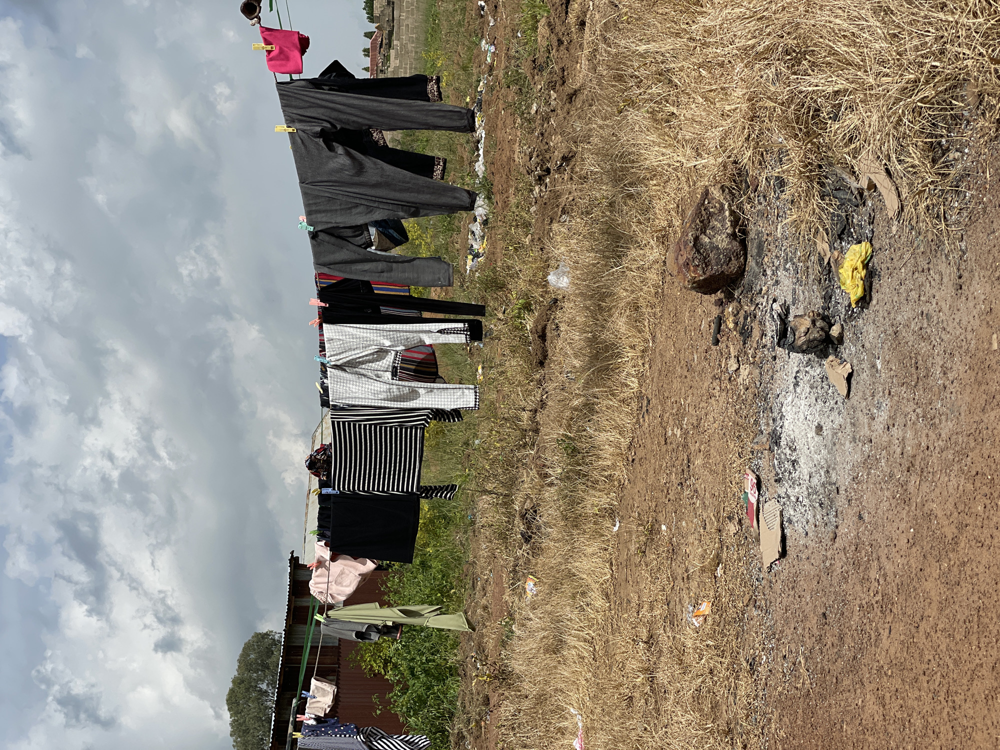 A clothesline stretches across the frame, laden with laundry. Dark grey pants hang prominently, alongside striped shirts and other garments. A bright pink bag is clipped to the line near the top. The clothes are hung on a wire using clothespins. In the background, a few small buildings with corrugated metal roofs are visible. To the right of the clothesline, the ground is covered in dry, tall grass and dirt. Some scattered debris, including rocks and what appears to be ash or burnt material, is present on the ground. The sky above is partly cloudy, with patches of white and grey clouds against a lighter sky. The overall scene suggests an outdoor domestic setting, likely in a rural or less developed area. The lighting indicates it is daytime. There is no visible text in the image.