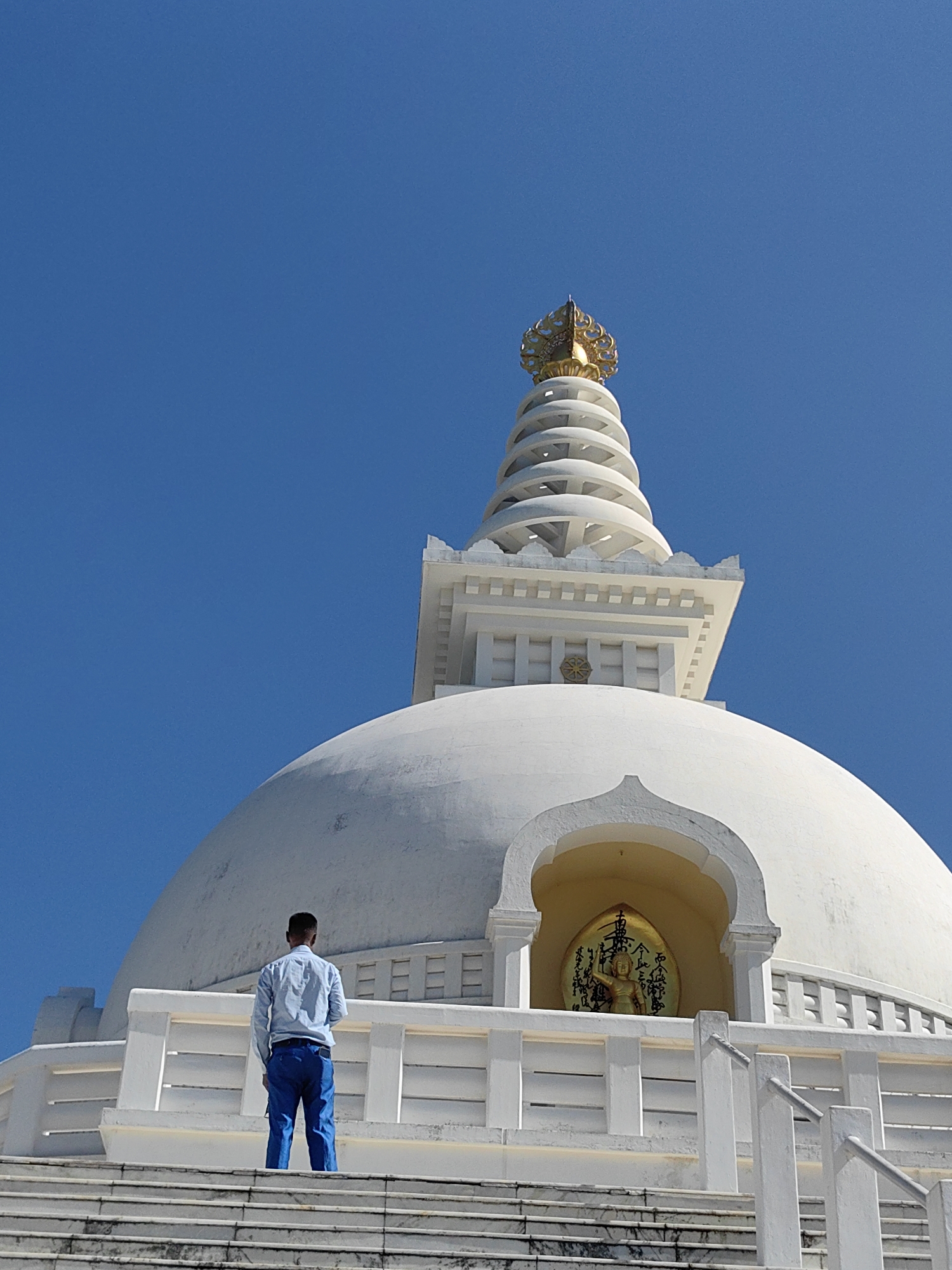 A man in a light blue button-down shirt and blue pants stands on white stairs looking at a white stupa against a clear blue sky. The stupa has a large dome and a tiered spire topped with a golden finial. A small alcove in the stupa's dome features a golden Buddha statue against a gold background with black calligraphy. White railings and decorative elements are visible on the stupa's exterior. The man is positioned on the stairs, facing away from the camera and towards the stupa. The scene appears to be in a public space, likely a religious or historical site.