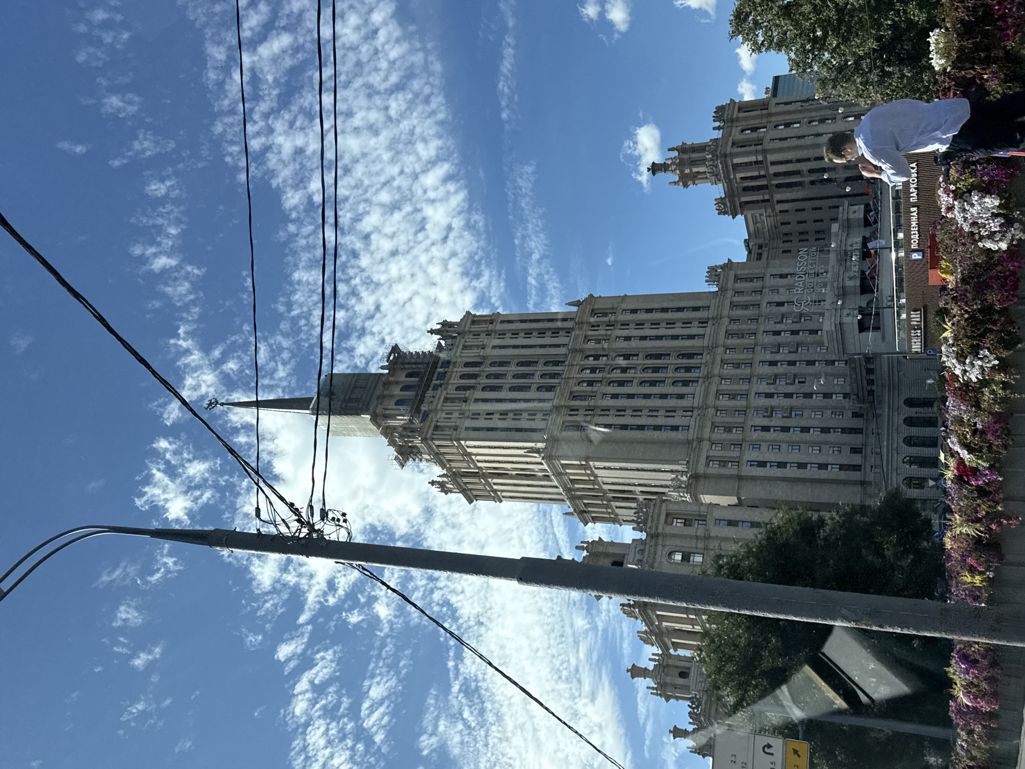 The image captures a view of the iconic Stalinist skyscraper, Radisson Collection Hotel, Moscow, also known as the Ukraina Hotel, situated in Moscow, Russia. The imposing building, characterized by its distinctive spired architecture, dominates the frame against a backdrop of a bright blue sky dotted with fluffy white clouds. In the foreground, a tall lamppost with tangled wires is prominently positioned, partially obscuring the view of the hotel. Below the hotel, a sign for "Radisson Collection Hotel, Moscow" is visible, alongside what appears to be an entrance labeled "CONGRESS PARK" and a parking sign indicating "ПОДЗЕМНАЯ ПАРКОВКА" (underground parking). A man dressed in a light blue shirt and dark pants stands near the bottom right of the image, facing away from the camera, appearing to be taking a photograph or interacting with his phone. The foreground also features a vibrant display of colorful flowers, suggesting a well-maintained urban environment. The presence of overhead wires and the perspective from what seems to be a moving vehicle (indicated by reflections on a glass surface on the left) suggest the photograph was taken from a street or a bridge.