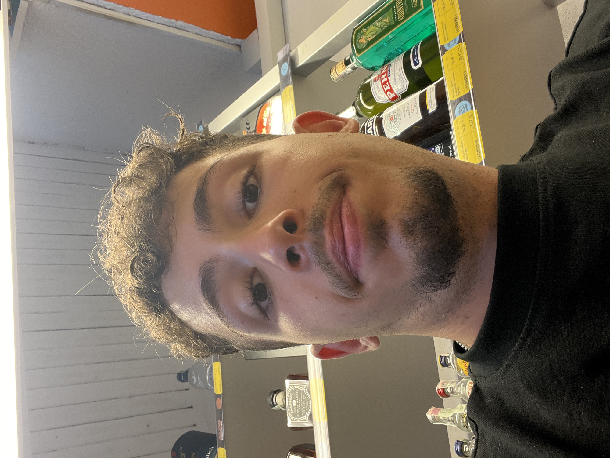 The image is a selfie taken by a young man with curly brown hair and a mustache and beard. He is wearing a black crew-neck sweatshirt and is positioned in front of shelves stocked with various bottles of alcohol. In the background, on the shelves, are bottles of Jack Daniel's Single Barrel, Pastis, Pernod, and Absinthe. There are also smaller bottles of spirits on a lower shelf. The lighting suggests an indoor environment, likely a liquor store or supermarket aisle. The man's head and shoulders occupy the majority of the frame, with the background shelves providing context. The overall impression is a casual, self-taken photograph within a retail setting. The image does not contain any discernible elements that would indicate the specific location within the city of Varna, Bulgaria.