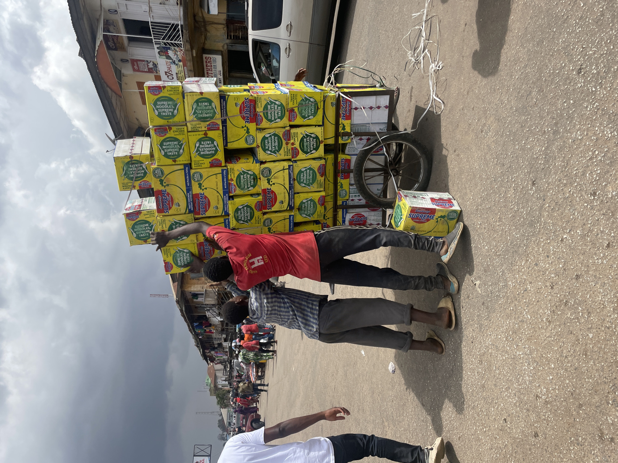 Two men are moving a large stack of "Supreme" brand instant noodles boxes loaded on a wheelbarrow on a street in Jos, Nigeria. The boxes are yellow with green and red labeling. One man wears a red shirt, while the other wears a checkered blue and white shirt. A third man in a white shirt is walking nearby. The street is paved and crowded, with buildings visible in the background, including a shop named "07SHUZE OTHERS, ORIES & GEM.TOOLS WAY. JOS." The sky is overcast.