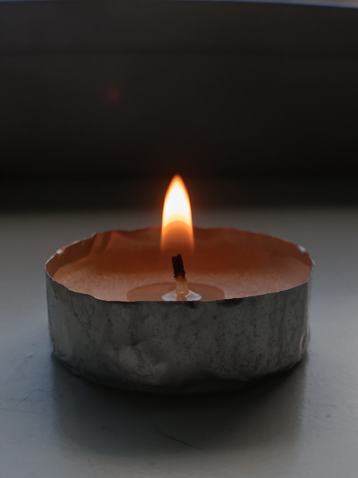 A close-up, eye-level shot shows a single lit tea light candle. The candle is housed in a silver metal tin with a slightly uneven rim. The wax inside the tin is a pale orange color. A bright, orange flame flickers at the top of a dark wick. The background is a dark, out-of-focus expanse, suggesting an indoor setting. The surface the candle rests on appears to be a light-colored, possibly reflective, surface with some subtle shadows. The scene is dimly lit, with the candle flame being the primary light source.