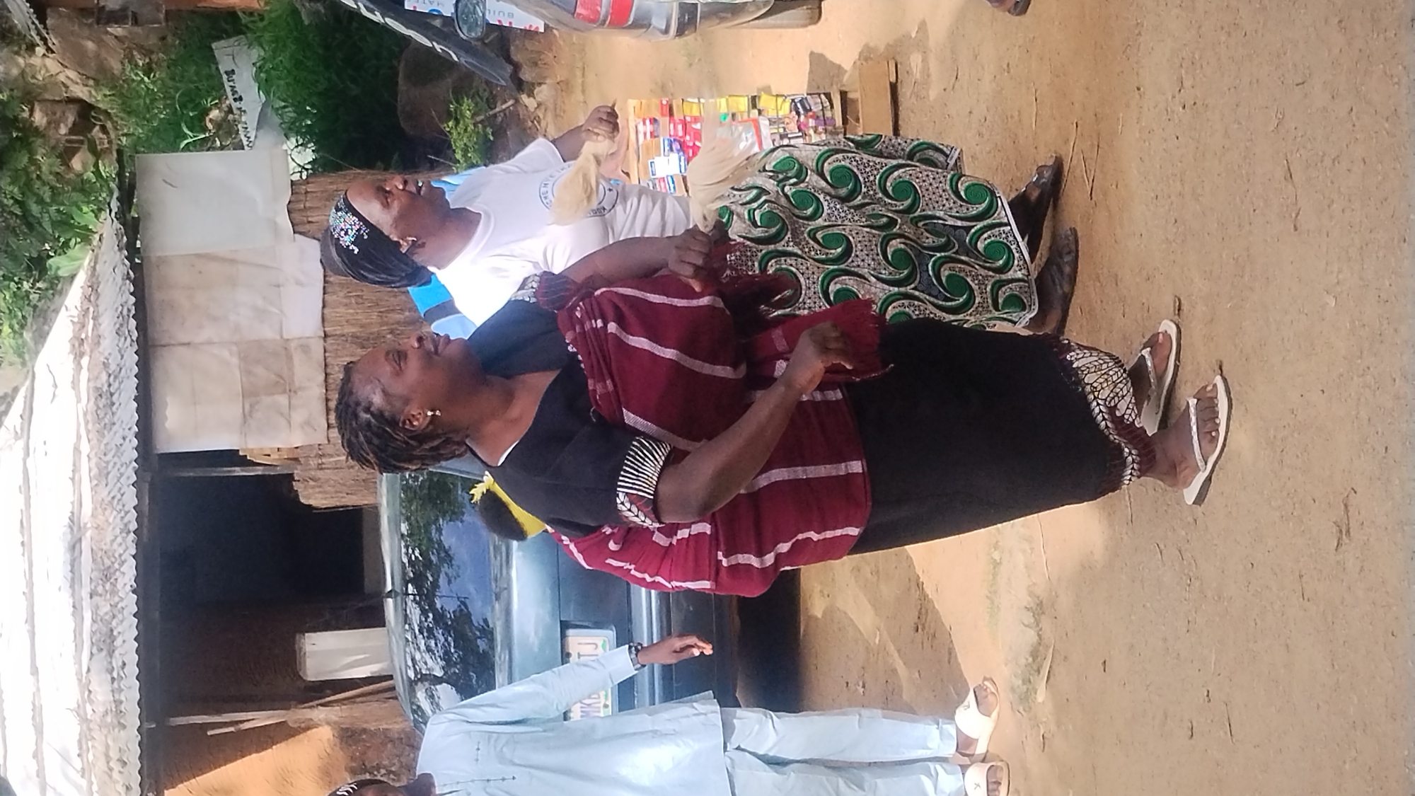 The image depicts a group of people in what appears to be a market or roadside stall setting in Jos, Nigeria. The scene is outdoors, with a sandy ground and some vegetation visible in the background. A corrugated metal roof structure is present on the left, suggesting a makeshift stall or shelter.
In the foreground, three individuals are prominently featured. The person closest to the camera is wearing a light blue traditional outfit and white flip-flops. Their hands are extended forward. To their right, another person is seated, wearing a dark outfit with a maroon and white striped shawl draped over their shoulders. They are looking upwards and to the left, with a smile. This individual has braided hair and is wearing earrings and a bracelet.
Further back and to the left, another person is also seated, wearing a white t-shirt and a patterned fabric on their lap. This individual is also looking upwards and to the left, with their mouth open in what seems to be laughter or animated speech. They have braided hair and are wearing a beaded headband.
In the background, a vehicle, possibly a car, is partially visible. Behind the seated individuals, a stall with various colorful items, possibly packaged goods or snacks, is displayed on a table or stand. The lighting suggests it is daytime, likely morning or afternoon, with mild weather. The overall mood appears cheerful and social, with the individuals engaged in lively conversation or interaction. No visible text is discernible from the image, other than what might be on the product packaging in the background.