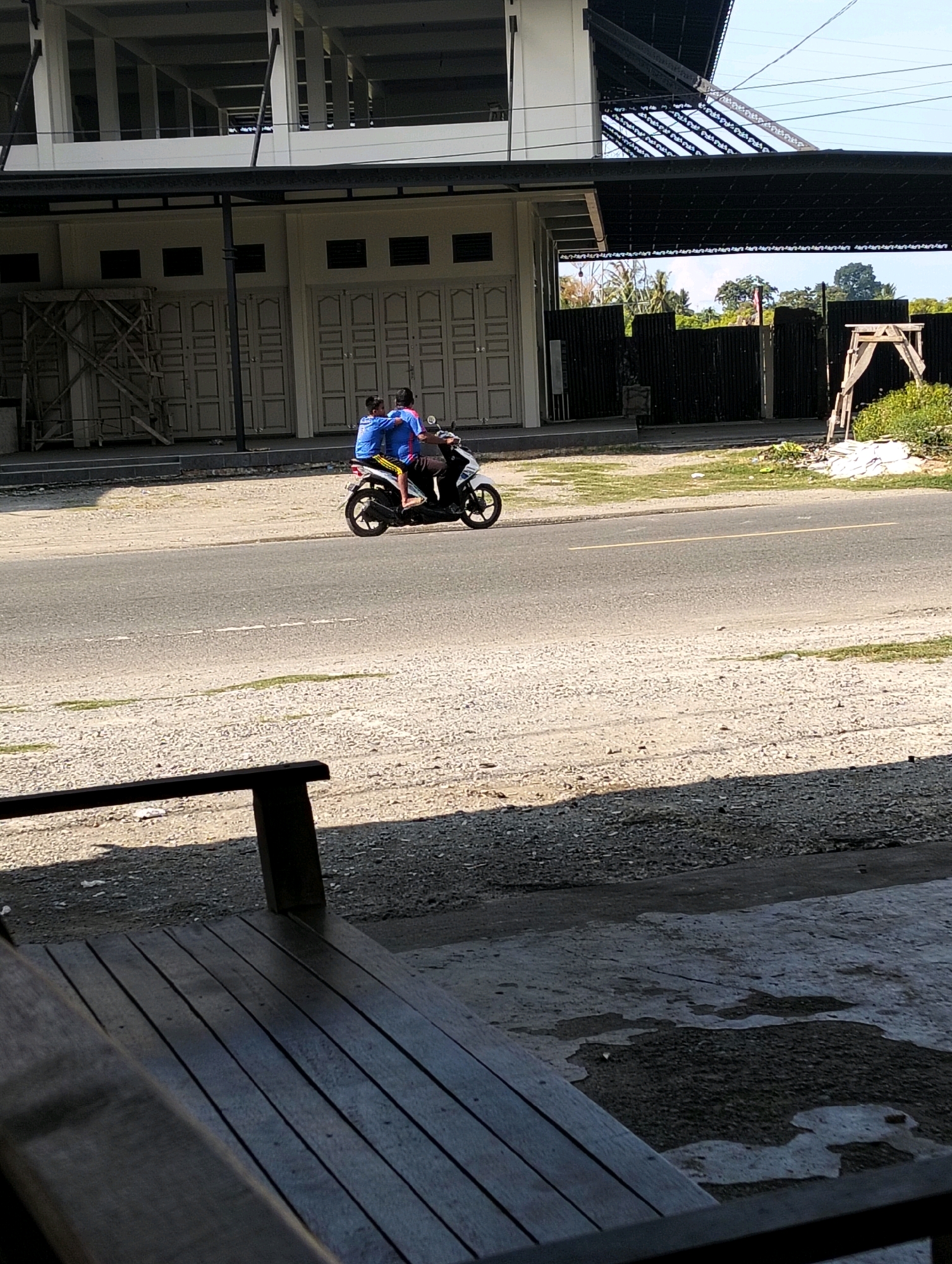 Two individuals are riding a white scooter on a paved road in Grong-Grong, Indonesia. The person in front is wearing a blue shirt, and the person behind is wearing a blue and pink shirt. The scooter has yellow and black accents on the side. They are traveling from left to right in the frame. The background features a building with white walls and dark doors, some scaffolding on the left, and a black corrugated metal fence with greenery behind it. The road is flanked by a gravel shoulder. In the foreground, a wooden bench is partially visible, casting a shadow. The sky is clear and blue.