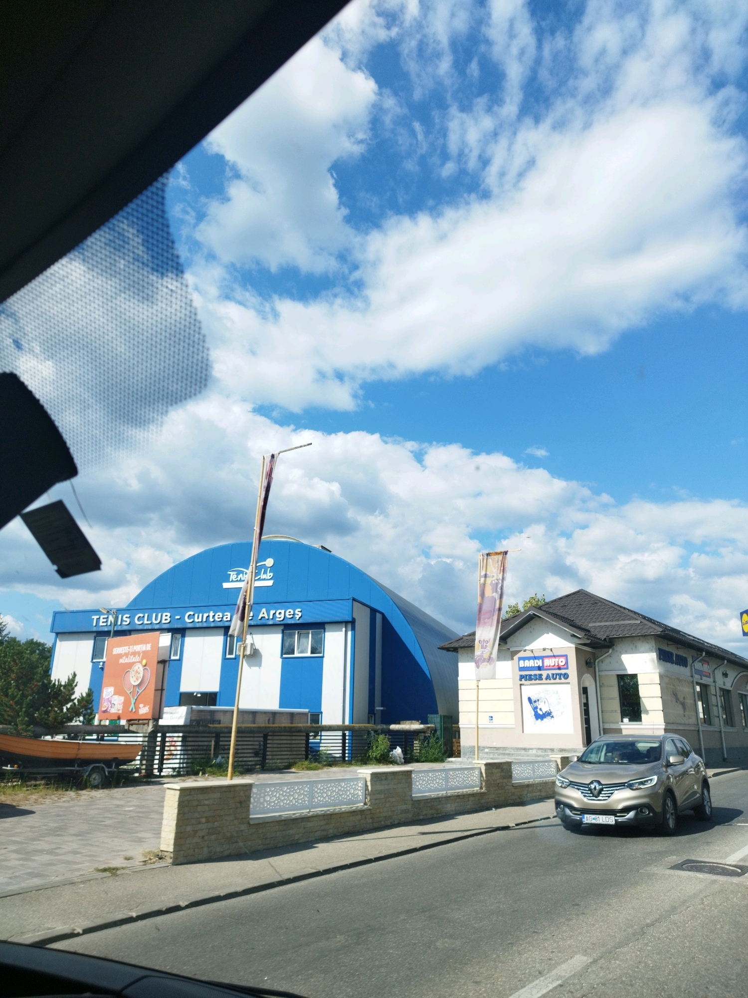 The image captures a street scene in Curtea de Argeș, Romania, on a bright, cloudy day. In the mid-ground, a large, blue and white arched building with "TENIS CLUB - Curtea de Argeș" written on its facade is prominent. To the right of the tennis club is a smaller, light-colored building with a dark roof, housing a business called "BARDI AUTO - PIESE AUTO". A modern, silver Renault car with license plate AG 81 LDS is driving on the road in front of the auto parts store. A wooden boat on a trailer is parked to the left of the tennis club building. The sky is a vibrant blue with scattered white clouds. The foreground shows a section of a paved road and a sidewalk with a decorative stone wall. The image appears to be taken from inside a vehicle, looking out through the windshield, as indicated by the dark gray area at the top left, likely part of the car's interior and the windshield's tinting.