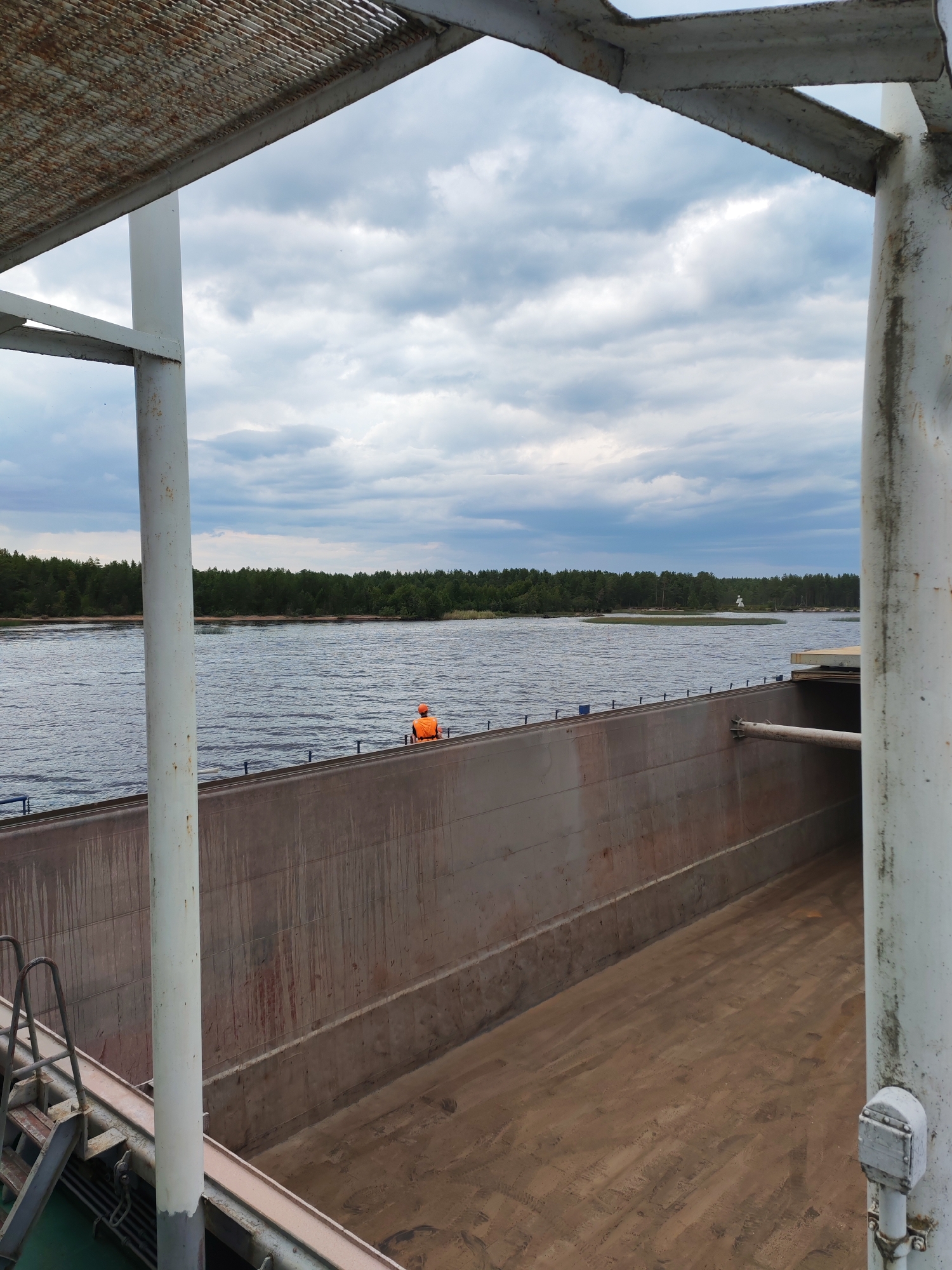 This is a photograph of a barge on a body of water, likely a river or canal, with a forested shoreline in the background. The image is taken from within the barge, looking out towards the water and the distant land. The barge itself appears to be an open-deck cargo vessel, with high, grey walls. The deck of the barge in the foreground is covered in sand or loose material. A metal ladder is visible on the left side of the barge. In the middle distance, a person wearing an orange safety vest is standing on the edge of the barge, looking out at the water. The water is dark and appears to be choppy. The shoreline is lined with dense green trees. The sky is cloudy with patches of blue showing through. The overall impression is one of an industrial waterway scene, possibly in an area with significant cargo transport.