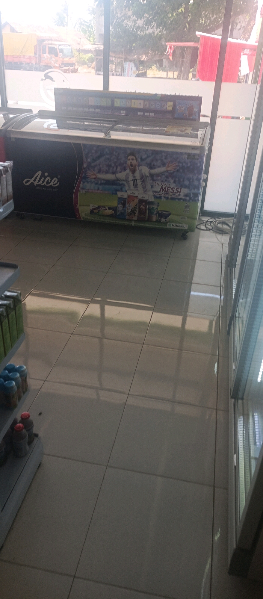 The image shows the exterior of a shop in Lamongan, Indonesia, with a large freezer decorated with a picture of Lionel Messi. The freezer is black with a red trim and features various Aice brand products. The shop appears to be located on a street, as a truck is visible in the background. The ground is tiled, and there are shelves stocked with bottles on the left side of the image. The overall impression is that of a small retail establishment in a somewhat rural or suburban setting.