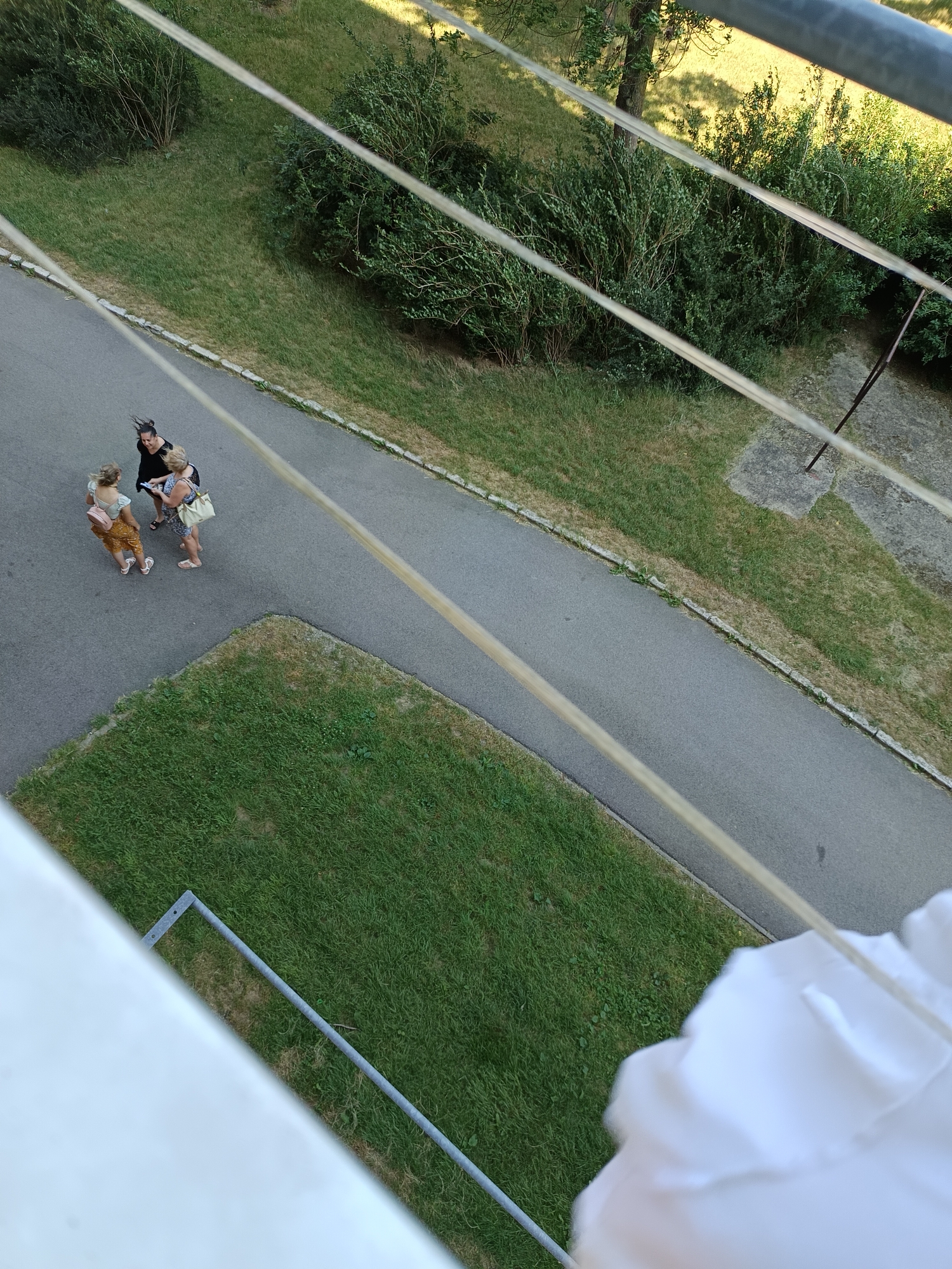 An aerial view captures three individuals standing on a paved pathway adjacent to a grassy area in Košice, Slovakia. Two women are positioned on the left side of the path, facing towards each other. The woman on the far left, wearing a light-colored top and orange skirt with sandals, holds a small pink bag. To her right, a woman in a black top and dark skirt engages in conversation. A third woman, on the right, wears a floral dress and carries a light-colored bag over her shoulder. Behind them, lush green bushes and trees line the pathway. The foreground is partially obscured by a white fabric object and metallic railing.