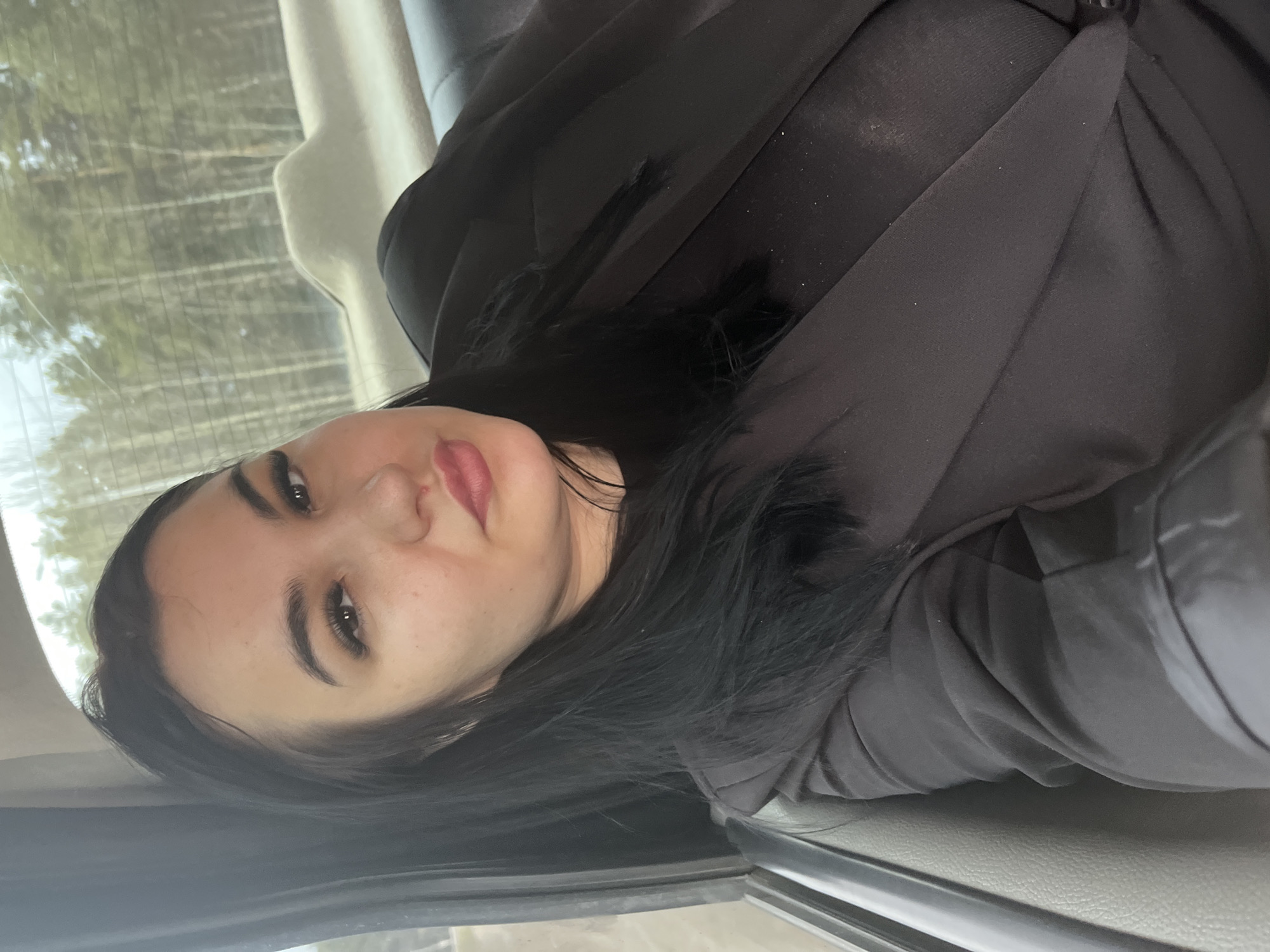 A woman with dark hair is sitting in a car. The background shows trees seen through the car window. The exact location in Chaikovsky, Russia, cannot be determined from this image.