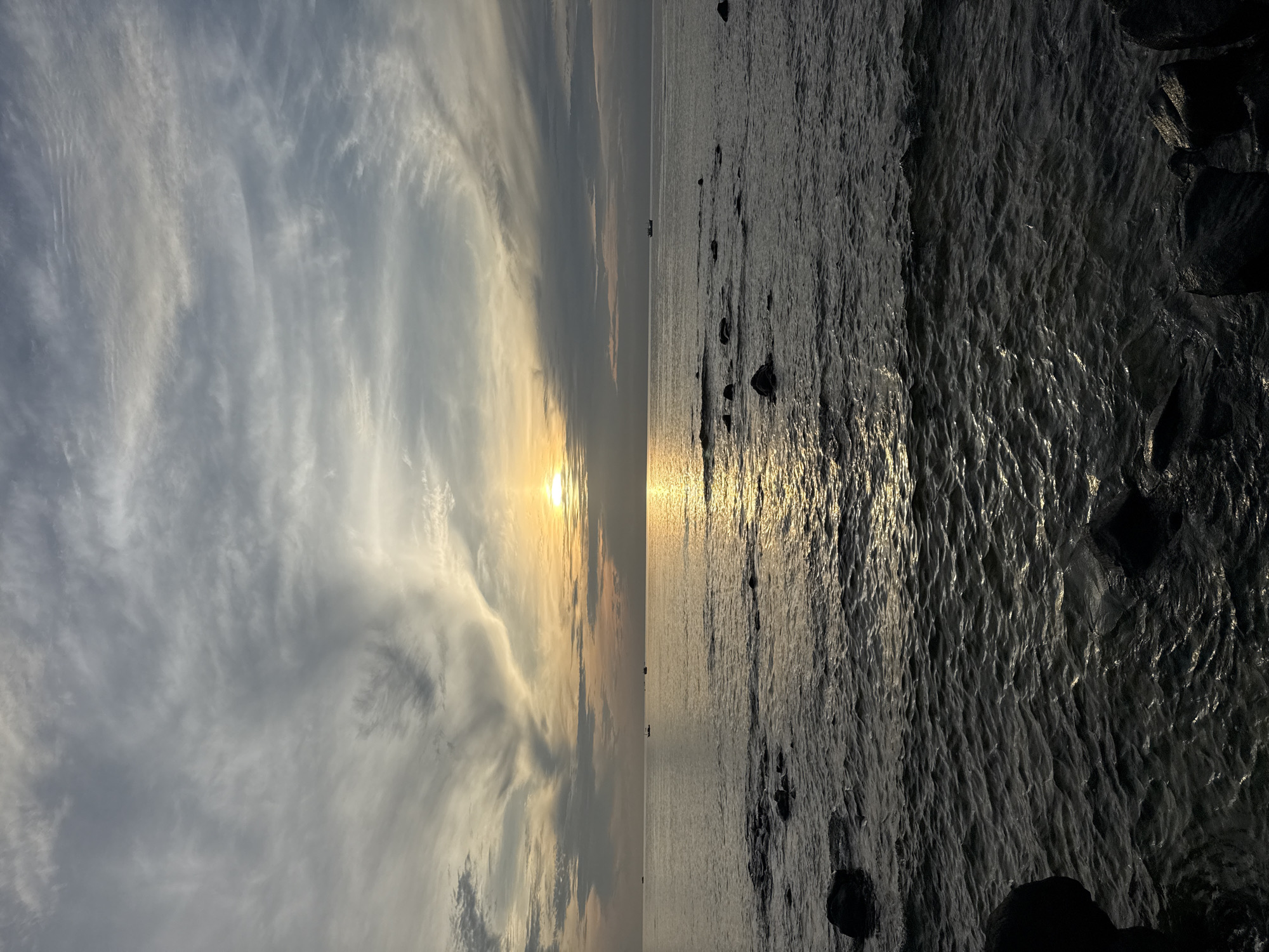 An outdoor, eye-level, medium shot captures the tranquil sea and sky of Lý Sơn Special Administrative Region, Vietnam at sunset. The sun, a bright orb, is positioned slightly right of center in the frame, casting a golden, shimmering path across the dark, rippling water. Wispy, light-colored clouds form intricate patterns against the grey sky, with a hint of orange and yellow near the horizon. Several small boats are visible in the distance, dotting the seascape. The foreground is occupied by dark, wet rocks partially submerged in the water, adding texture and depth to the scene. The overall atmosphere is serene and peaceful, with the natural beauty of the coastal landscape being the central focus.