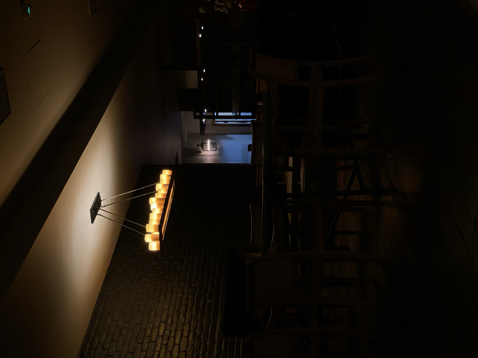 The image depicts the interior of a dimly lit restaurant in Stepantsminda, Georgia. A rectangular chandelier with multiple faux candles hangs suspended above a table setting. The walls are made of dark brick, and wooden chairs surround the table. In the background, another light fixture is visible on the wall, and shelves with glassware can be seen. The overall atmosphere is dark and intimate, with the light from the chandelier casting a warm glow on the surrounding area. The scene appears to be unoccupied.