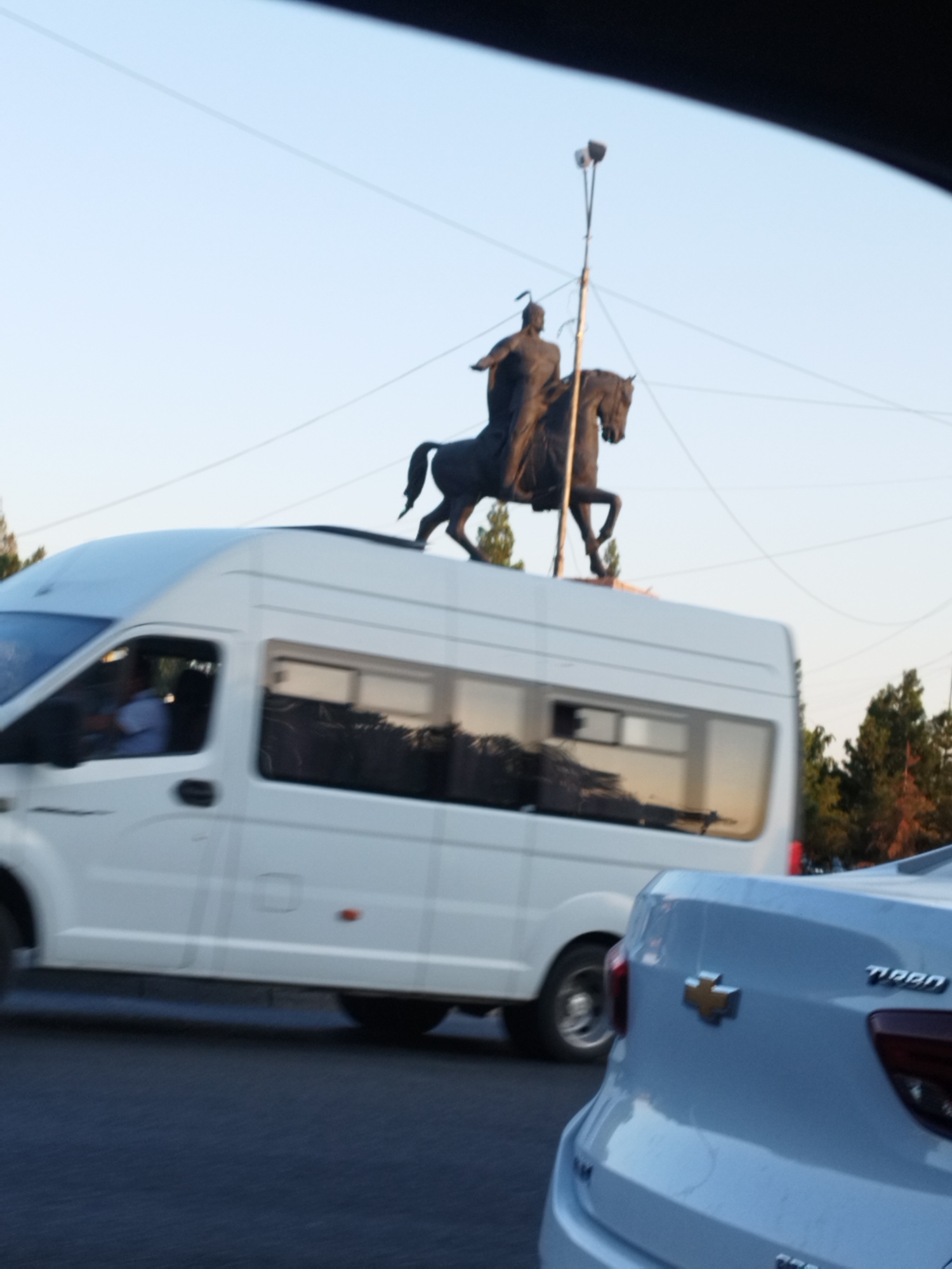 A bronze equestrian statue of a man in armor, possibly a warrior or historical figure, stands against a clear sky in Shymkent, Kazakhstan. The statue is located above street level, potentially on a monument or elevated platform. Below the statue, a white commercial van, likely a GAZelle, is driving on a road. Its windows reflect the surroundings, including trees and the sky. To the right of the van, the rear of a light blue Chevrolet sedan is visible. The sedan has a Chevrolet emblem on its trunk and the word "TIBA" or a similar inscription. The scene appears to be a street or intersection in the city, with power lines and trees also present in the background. The lighting suggests it is daytime, possibly late afternoon or early morning due to the soft light.
