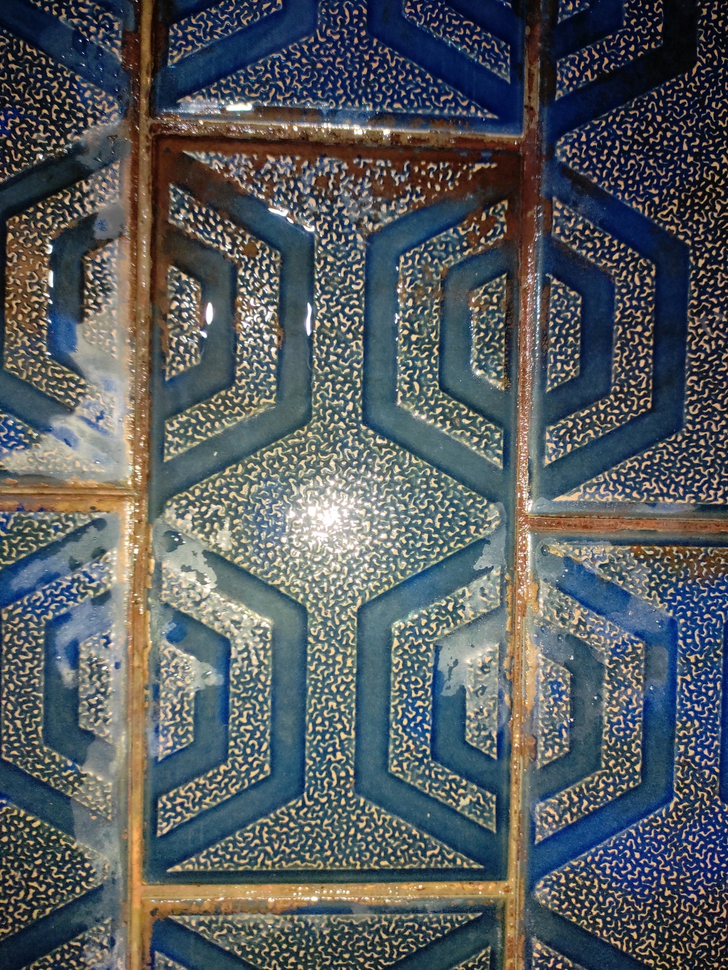 This is a close-up, eye-level photograph of a tiled surface. The tiles are square and appear to be made of ceramic with a glossy finish. The dominant color is a deep blue, with a textured, speckled pattern in a lighter beige or cream color. Overlaid on this textured surface are raised geometric patterns in a darker blue. These geometric shapes form a hexagonal or honeycomb-like design that is repeated across the tiles. The grout lines between the tiles are visible and appear to be aged and slightly discolored, with some areas showing signs of rust or dirt accumulation. There are also small droplets of water or moisture on the surface of some tiles, reflecting the light. The lighting in the image is somewhat harsh, causing bright reflections on the glossy surfaces of the tiles. The overall impression is of an older, possibly outdoor, tiled surface that has experienced some wear and tear.