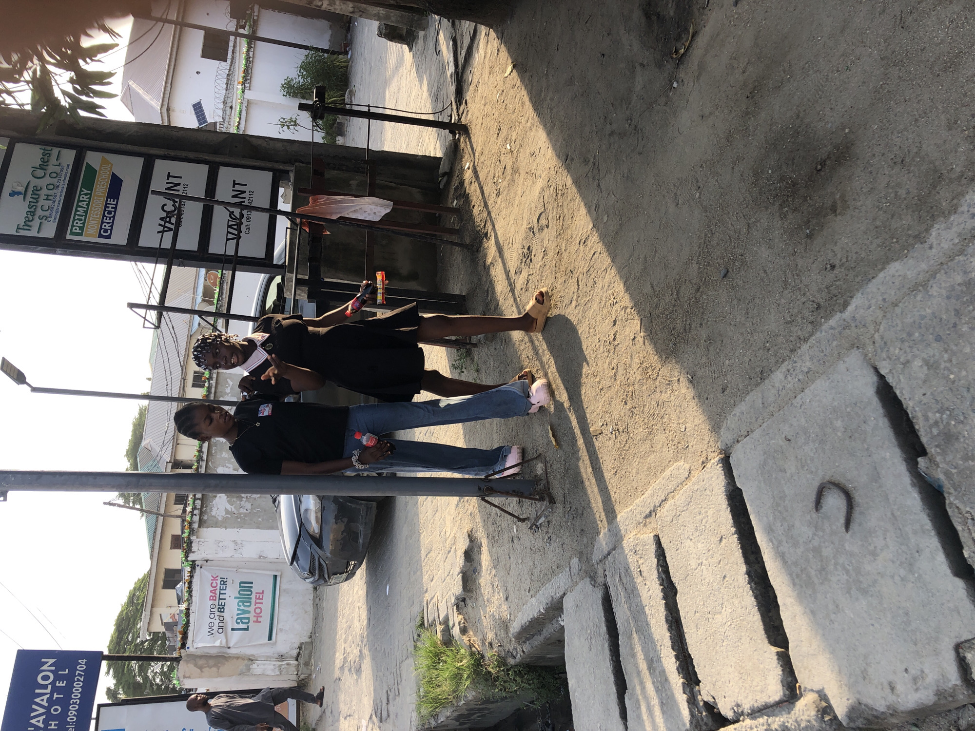 The image depicts an outdoor scene in Ado, Nigeria. Two women are visible. One wears a black top and blue jeans with pink sneakers and holds a red bottle. The other woman wears a black dress and sandals. The women are standing near various signs advertising local businesses. These signs include Avalon Hotel, Lavalon Hotel, and Treasure Chest School. Adjacent to Treasure Chest School is a sign saying “Vacant.” A gray car is parked nearby. The ground is dirt and concrete. A concrete drainage ditch is located on the side of the image. Another individual is seen walking away from the camera near the Avalon Hotel sign.