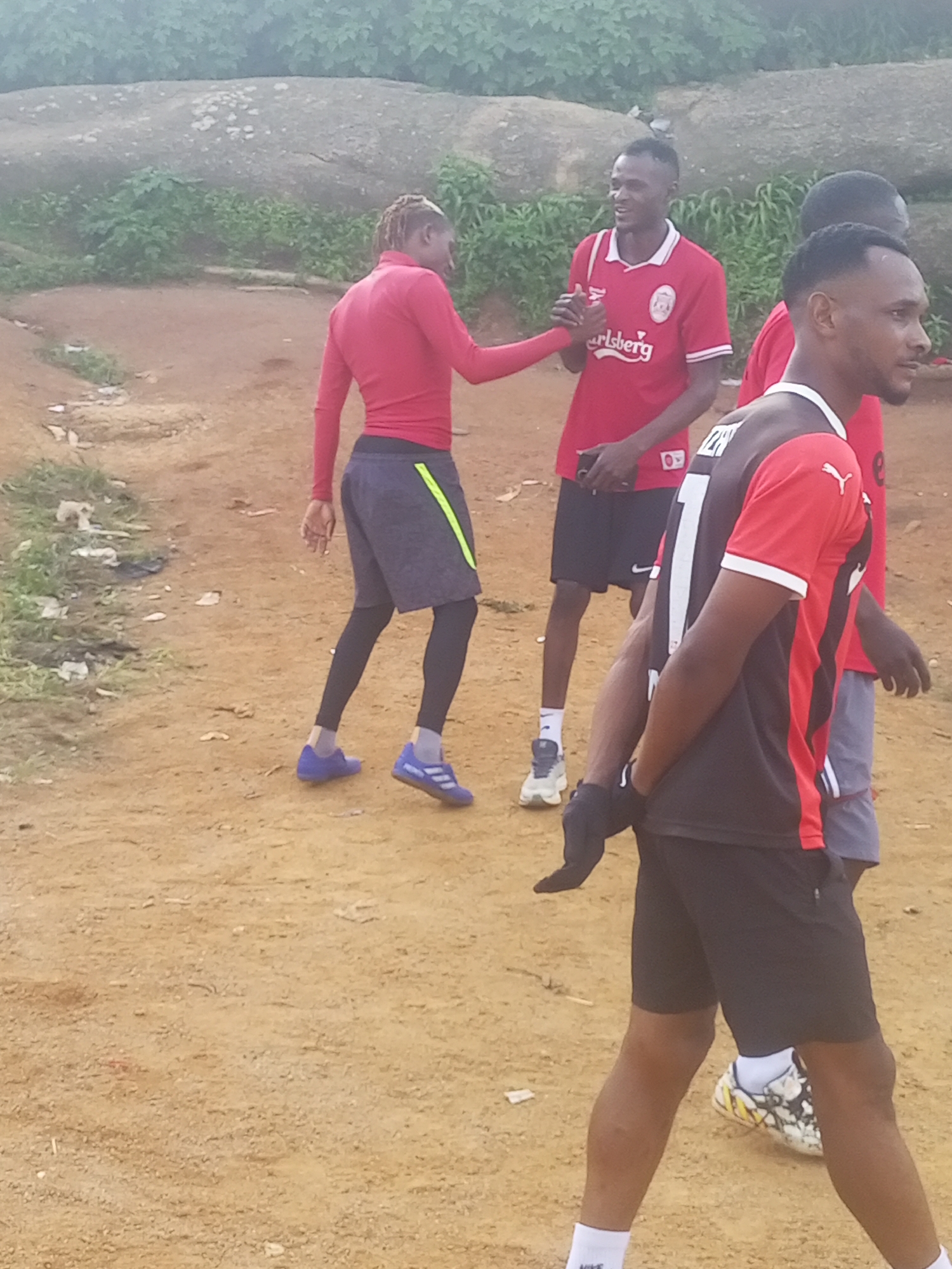 In the city of Jos, Nigeria, four young men are gathered outdoors on a dirt ground with scattered rocks and vegetation in the background. Three of the men are wearing sportswear, with two in red shirts and shorts/leggings, and one in a black and red jersey. The fourth man is partially visible, wearing a red shirt. One man in the red shirt is seen shaking hands with another man in a red shirt. The atmosphere appears casual and relaxed, possibly before or after a sporting activity.