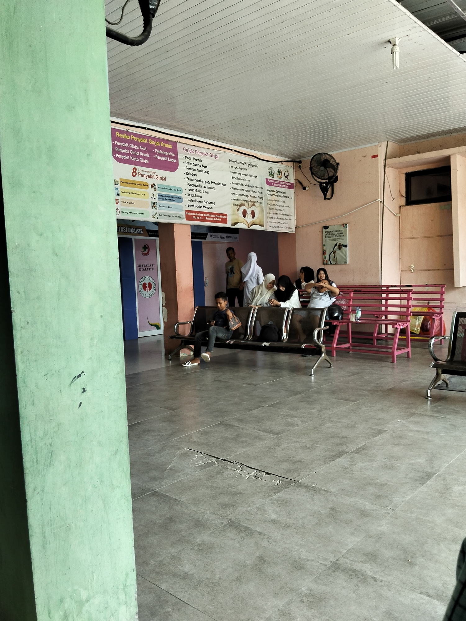 This is a medium-angle, full shot taken in what appears to be the waiting area of a medical facility, likely a hospital or clinic, given the presence of "INSTALASI DIALISIS" (Dialysis Installation) signage. The overall scene is somewhat dated, with a pale pink wall and a white, paneled ceiling. Fluorescent lights are visible, and a dark industrial fan is mounted on the wall. The floor is tiled in large, gray squares, with some visible cracks.
Several people are present. In the foreground, a young person sits alone on a dark-colored bench, looking down at their phone. Further back, a group of women are seated on a pink bench, some wearing headscarves. Another person, a man in a yellow shirt, stands near a doorway in the background.
Prominently displayed on the wall is a large informational poster with Indonesian text, detailing risks, symptoms, and prevention of kidney disease ("Resiko Penyakit Ginjal Kronis," "Gejala Penyakit Ginjal," "Cara Mencegah Penyakit Ginjal"). Another smaller poster, likely an advertisement or information about service usage, is also visible near the entrance. The presence of the dialysis installation sign suggests a connection to kidney treatment.
The lighting suggests it's daytime, possibly late morning or afternoon, with natural light potentially coming from unseen windows. The overall atmosphere seems quiet and expectant, typical of a waiting room. The location context provided indicates the scene is in Bireuen, Indonesia.