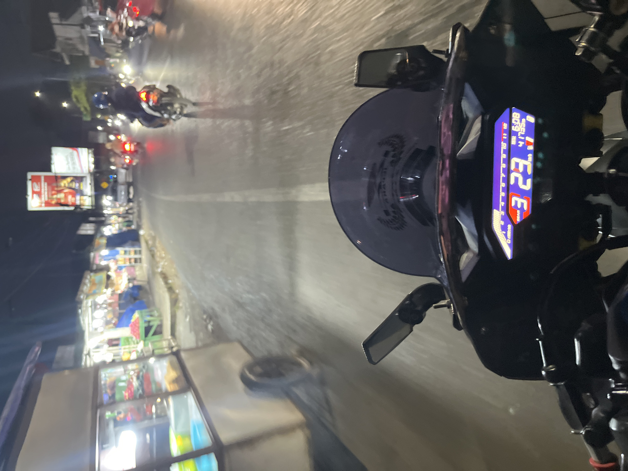This image is a first-person perspective shot from a motorcycle at night, on a street in Pecangaan, Indonesia. The motorcycle's digital dashboard is prominently displayed in the foreground, showing a speed of 23 km/h, a gear indicator of '3', a total time of 6:08, and a total distance of 41735 km. The dashboard is illuminated in a purple and blue hue. To the left, a food stall or cart with glass displays and visible food items is positioned alongside the street. The street itself is paved and appears wet, reflecting the lights from various sources. Several other motorcycles and vehicles are visible on the road, some with illuminated taillights. Buildings and streetlights line the side of the road, and advertising billboards are present in the background. The overall scene depicts a nighttime streetscape with active traffic and commercial activity.