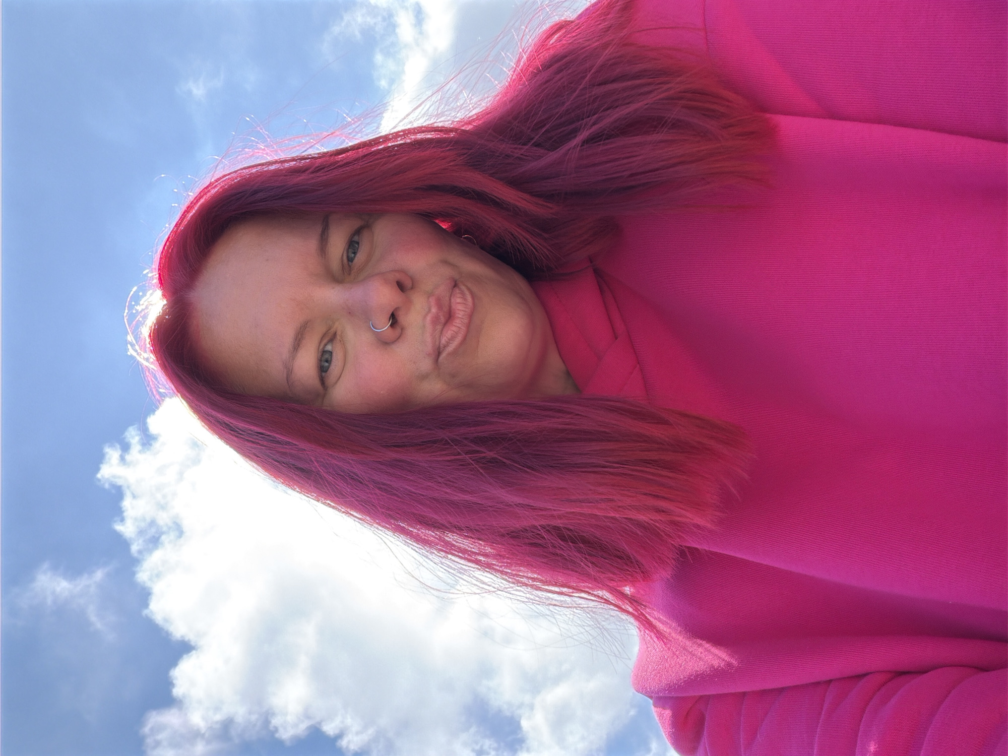 An extreme low-angle selfie captures a person against a bright blue sky dotted with scattered white clouds. The individual has vibrant, bright pink or magenta hair, which appears to be dyed. They are wearing a matching pink hooded sweatshirt or top. Their face is angled towards the camera, with a silver hoop nose ring visible. Their lips are pursed in a playful "duck face" expression, and their eyes are open, looking directly at the viewer. The sun is visible in the upper left, casting a bright backlight on their hair, creating a luminous glow around the edges. The person's left shoulder and arm are visible, clad in the pink garment. The scene depicts an outdoor setting under clear weather conditions, likely during daytime. There is no visible text. The location context for this image is Chaikovsky, Russia.