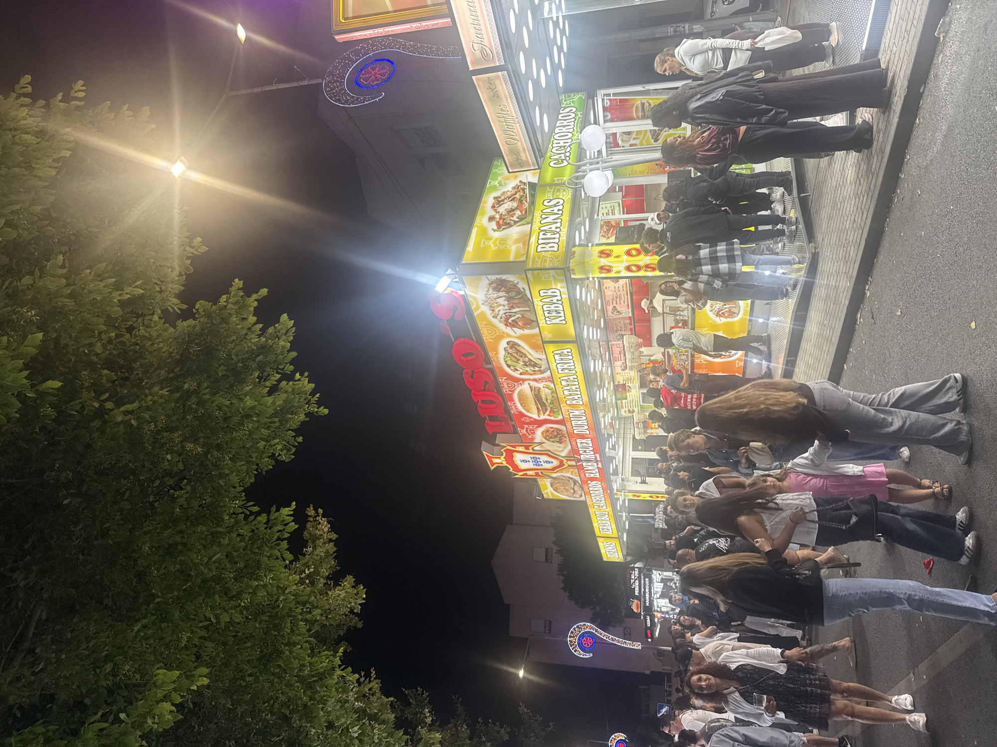 The image captures a lively nighttime scene at a food street market in Cascais, Portugal, likely during a local festival or event. A prominent food stall with bright yellow and red signage displays various offerings including "BIFANAS," "KEBAB," "DURUM," and "BATATA FRITA" (fries), alongside images of food items. Another stall to the right advertises "Waffles" and "Cachorros" (hot dogs). The stalls are illuminated by overhead lights, creating a festive atmosphere. A crowd of people, a mix of genders and ages, are gathered in front of the stalls, some queuing, others socializing. Their attire suggests casual evening wear. In the foreground, several individuals are captured mid-interaction, including a woman holding a drink and a group of young women conversing. The background shows buildings and trees, with decorative lights adding to the ambiance. The ground is paved with asphalt and cobblestones.