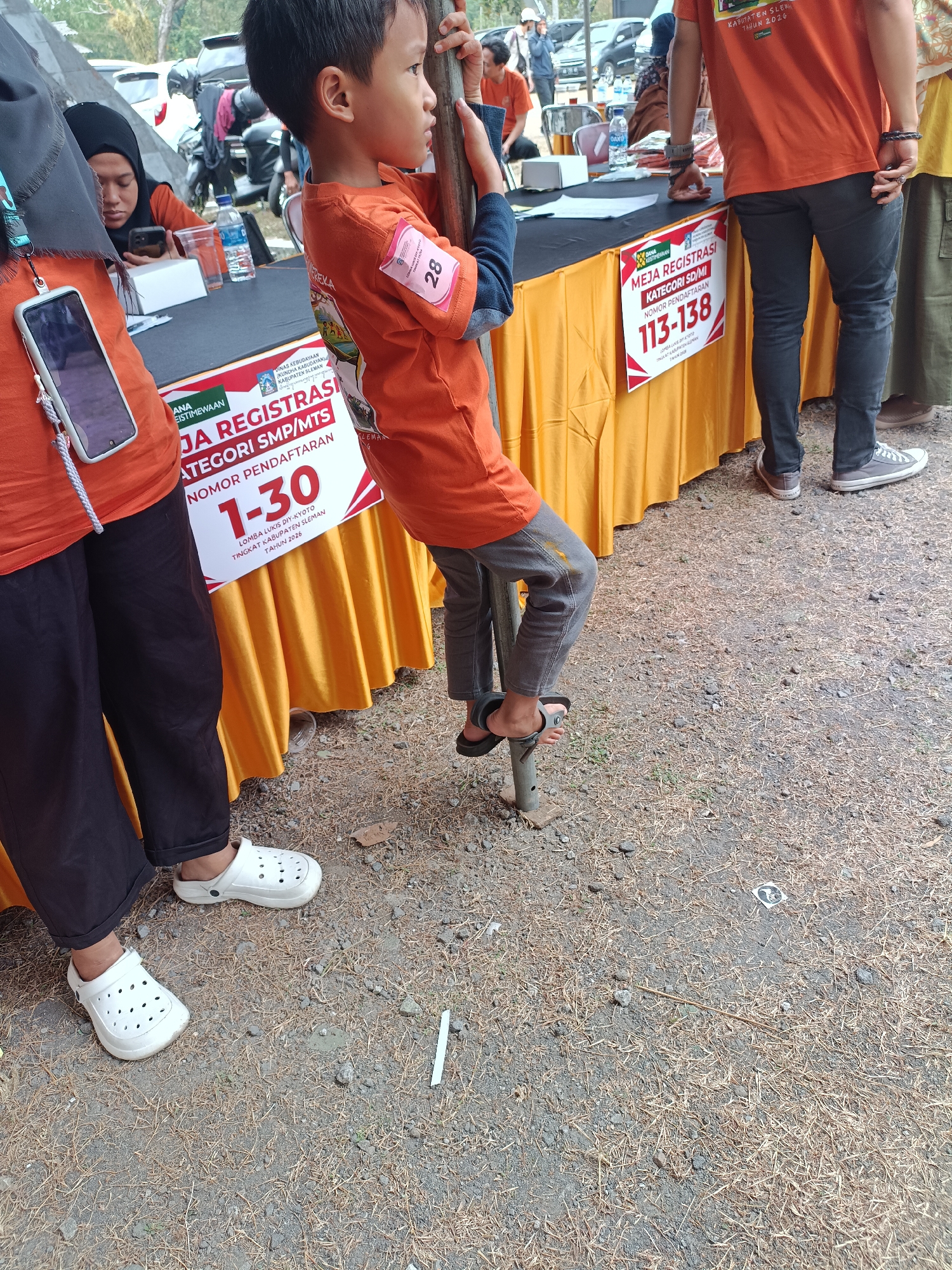 The image depicts an outdoor scene at a registration event in Hargobinangun, Indonesia. In the foreground, a young boy wearing an orange t-shirt and grey pants is holding onto a metal pole. He is looking to the right. To the left of the boy, two individuals are partially visible, also wearing orange t-shirts. One is wearing a black hijab. A table draped with an orange cloth is positioned behind them, displaying signage with text including "JA REGISTRASI KATEGORI SMP/MTS NOMOR PENDAFTARAN 1-30" and "MEJA REGISTRASI KATEGORI SD/MI NOMOR PENDAFTARAN 113-138". Several white boxes and a bottle of water are on the table. The ground is covered with dirt and dry grass. In the background, vehicles, including cars and motorcycles, are parked.