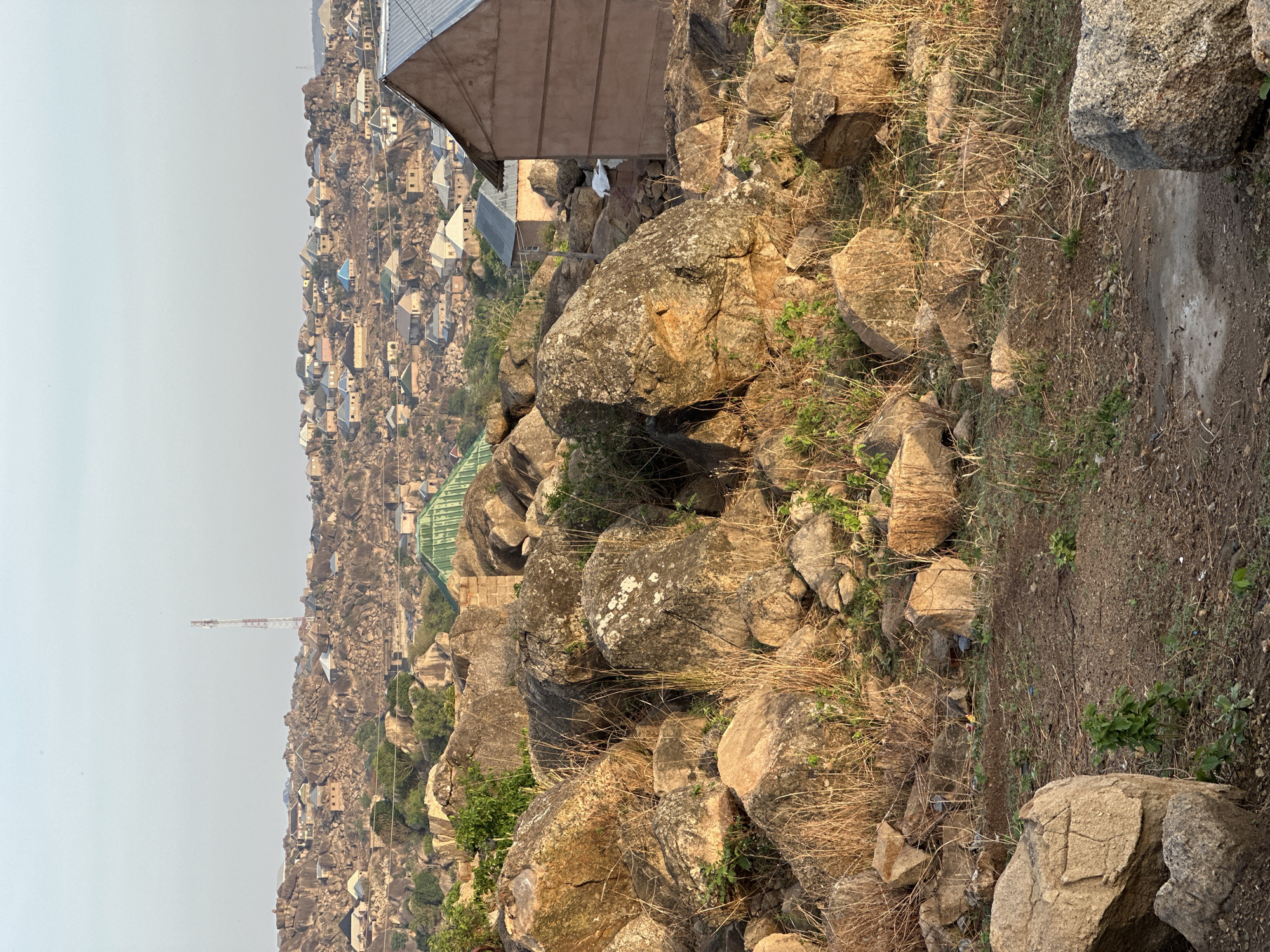 The image depicts a rocky, hillside scene in Jos, Nigeria. The foreground is dominated by large, lichen-covered boulders and dry vegetation. A narrow dirt path is visible. In the background, a densely populated cityscape of small buildings stretches into the hazy distance. A tall communication tower rises above the buildings. An artificial shelter is on the upper side of the frame.